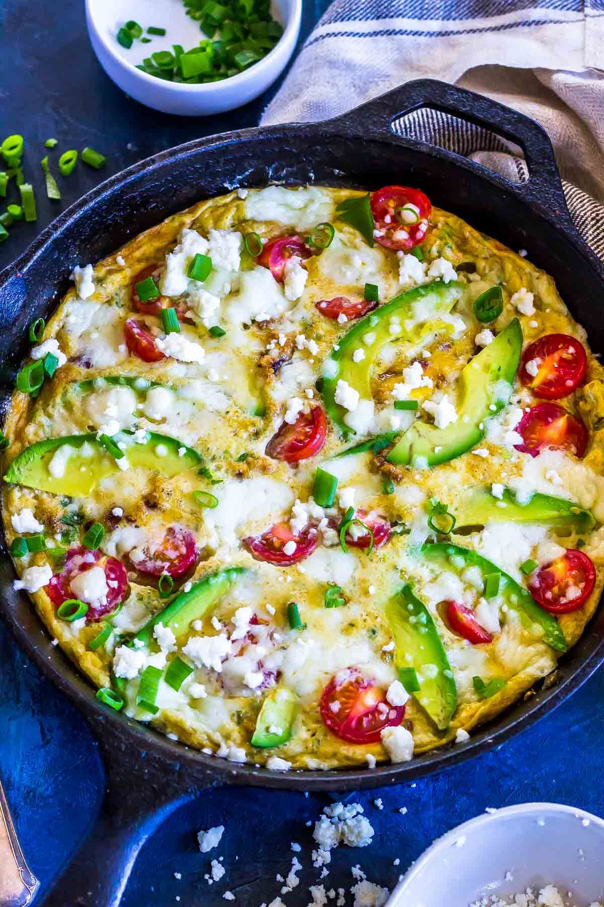 On a blue surface is a cast iron round pan with a frittata in it made of eggs, avocado slices, cherry tomatoes and with cheese crumbled over the top. A white dish with chopped scallions is just behind, next to a blue and white striped table cloth