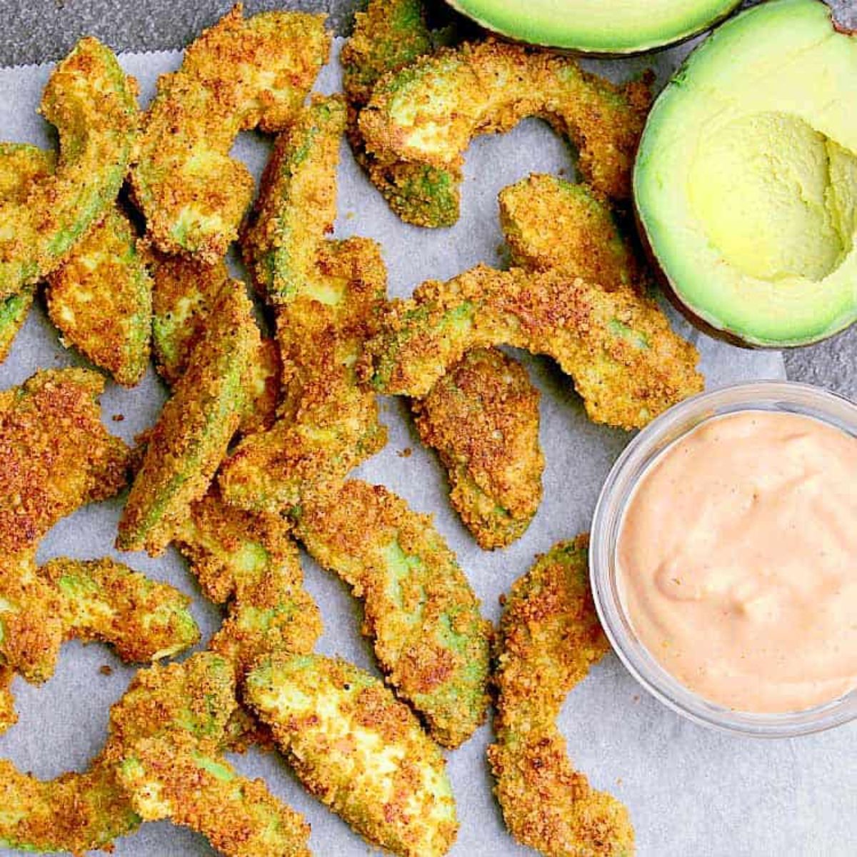 On a sheet of baking parchment are avocado fries. To the right is a glass pot of a pink dip and above are two avocado halves