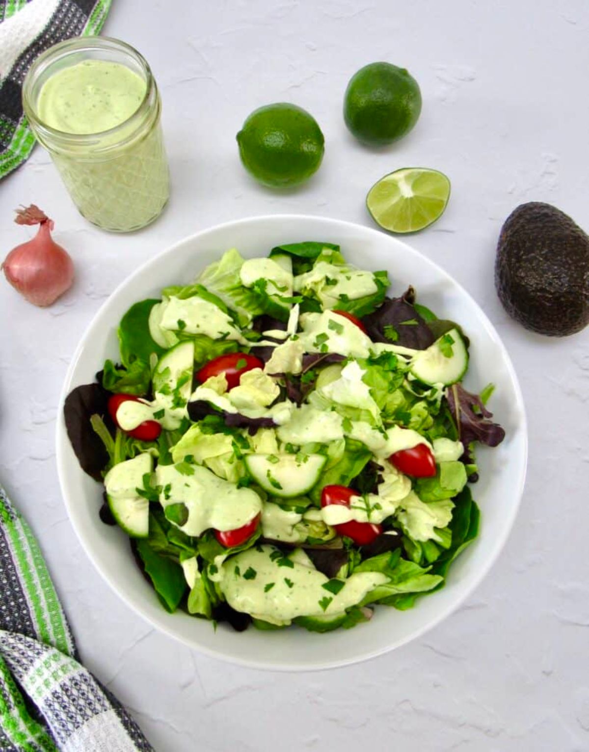 On a white marble countertop is a white round bowl with a green salad, drizzled with green dressing. Around the bowl is a glass jar of dressing, limes and lime wedges, a shallot, and an avocado