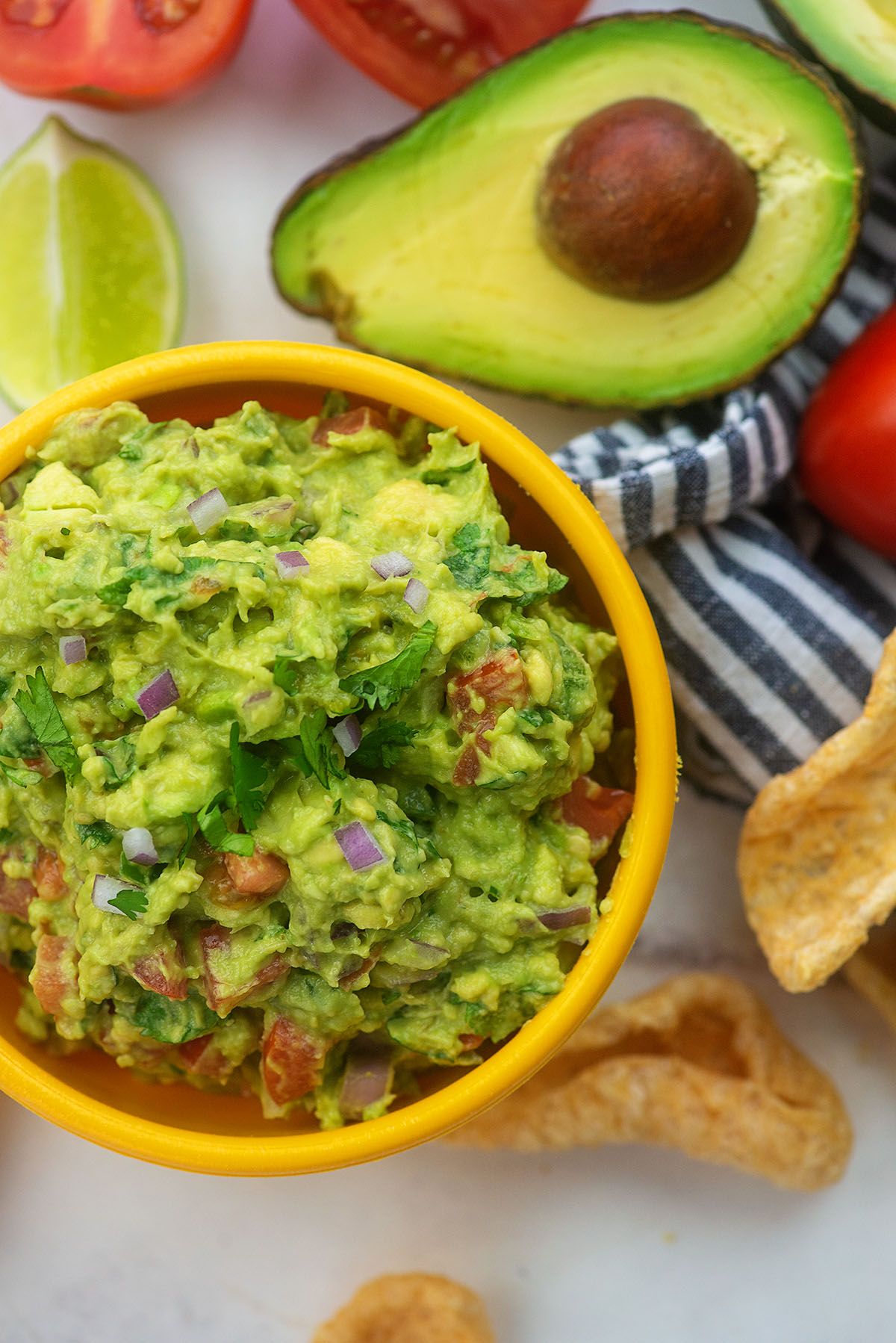 ON a white worktop is a small yellow bowl filled with guacamole. Next to it are some tomato halves, an avocado half, a lime wedge and some chips on a blue and white striped cloth