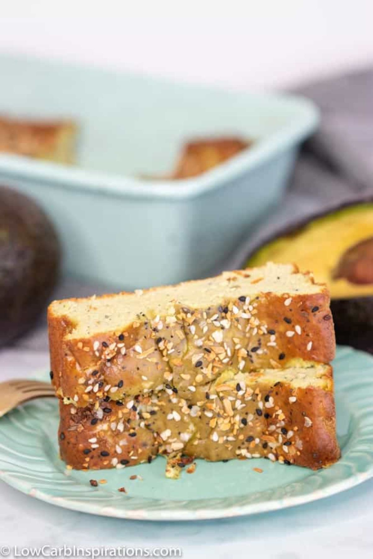 In the foreground on a light green plate are two thick slices of bread topped with seeds. Blurred in the background is a green baking tin, and some avocado halves