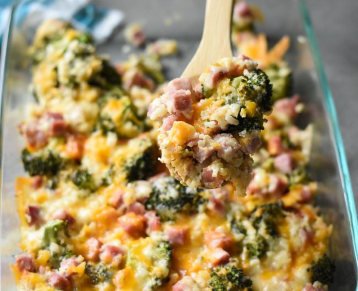 a blurred shot of a glass casserole dish filled with cheesey, broccoli and ham casserole. A wooden spoon lifts some of the casserole out