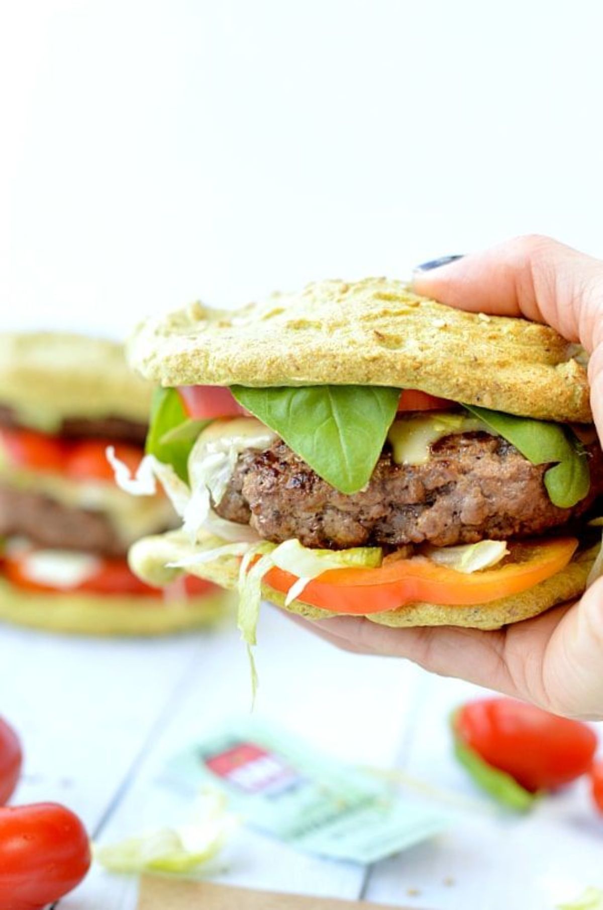 A hand holds a sandwich filled with green leaves, sliced tomatoes and a burger patty. In the background is another sandwich, and some cherry tomatoes