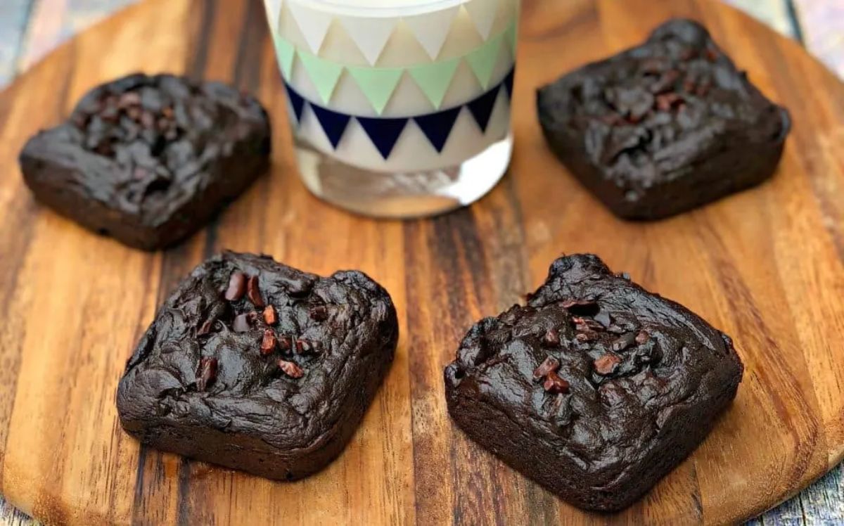 On a round wooden chopping board are 4 fudgy cocolate brownies, with a glass of milk in the middle