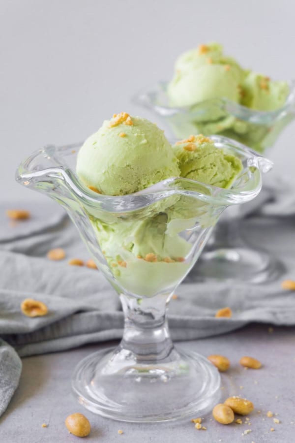 On a gay background with a gray cloth behind, are 2 ice cream glass dishes filled with 3 scoops of green ice cream and toppped with peanuts