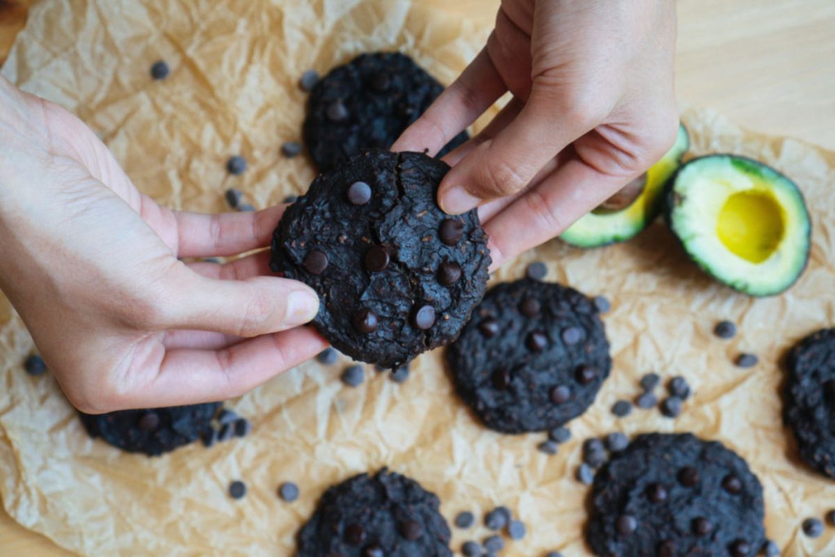 2 hands hold a round dark chocolate cookie above a sheet of baking parchment on which sit more cookies and 2 halves of an avocado