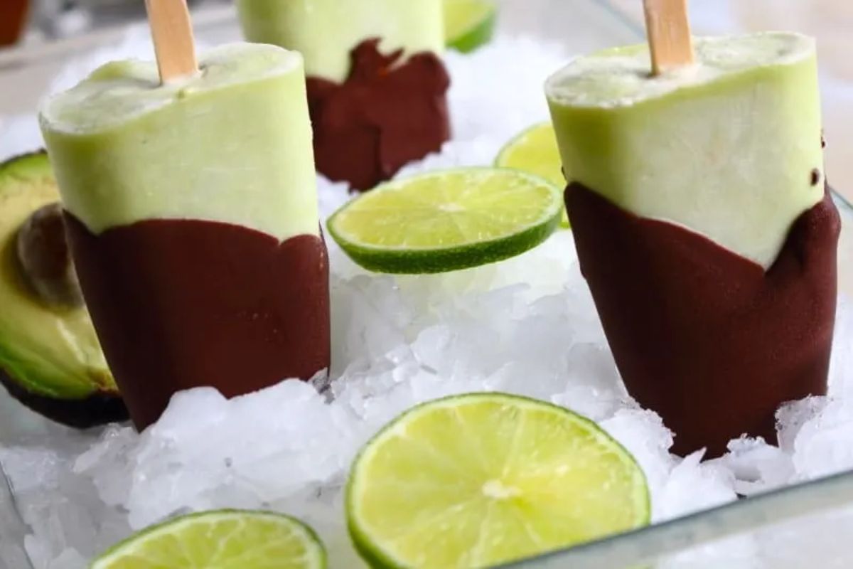 In an ice-filled tray are 3 green popsicles, with the tops dipped in chocolate. Dotted around are lime slices