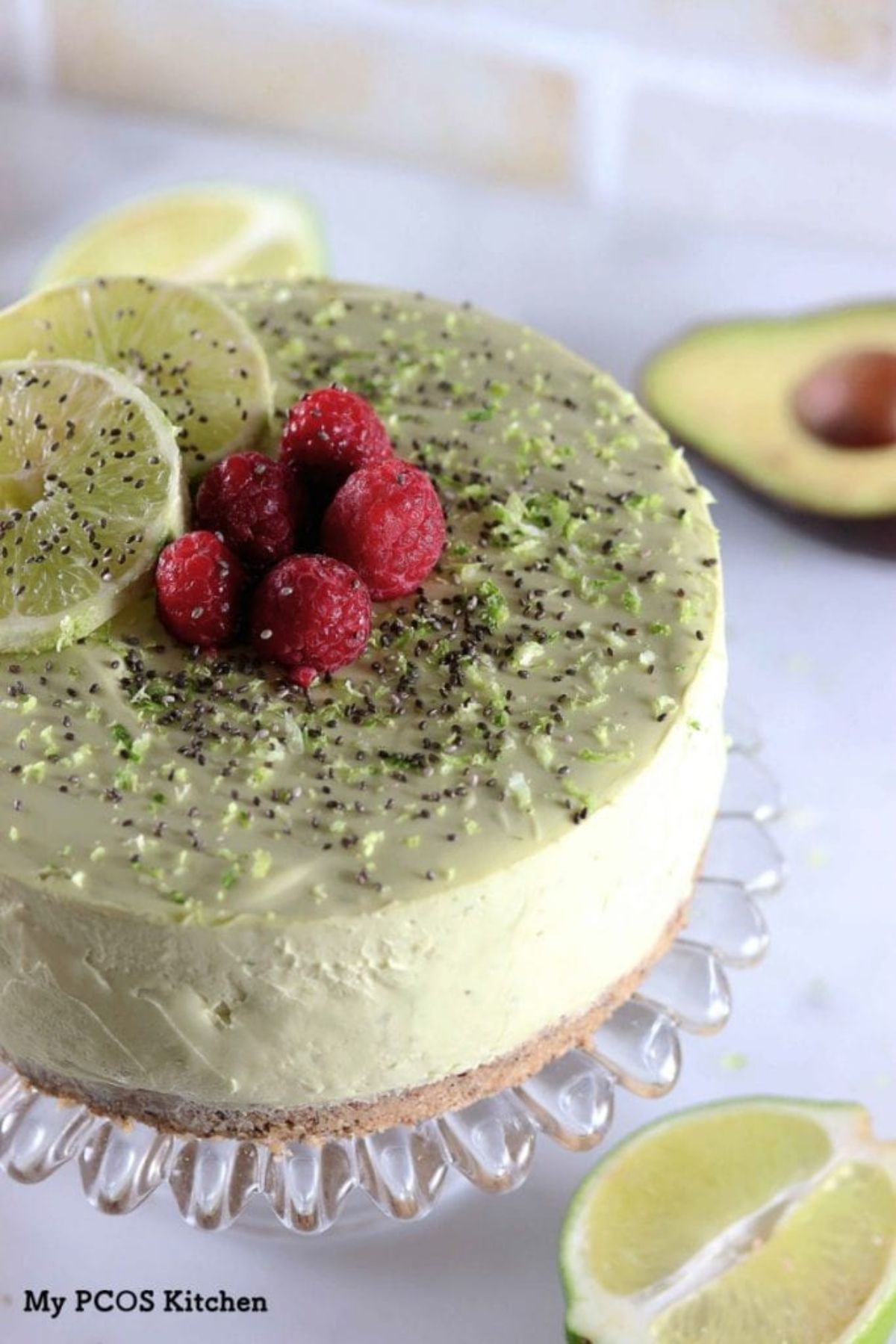 On a glass, fluted ckae stand is a light green cheesecake topped with lime zest, raspberries and lime slices. LIme wedges and an avocado half are off to the side