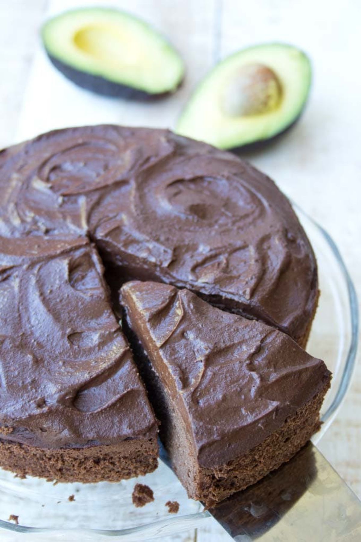 On a glass cake stand is a chocolate ckae with one slice cut out of it. Behind are 2 avocado halves