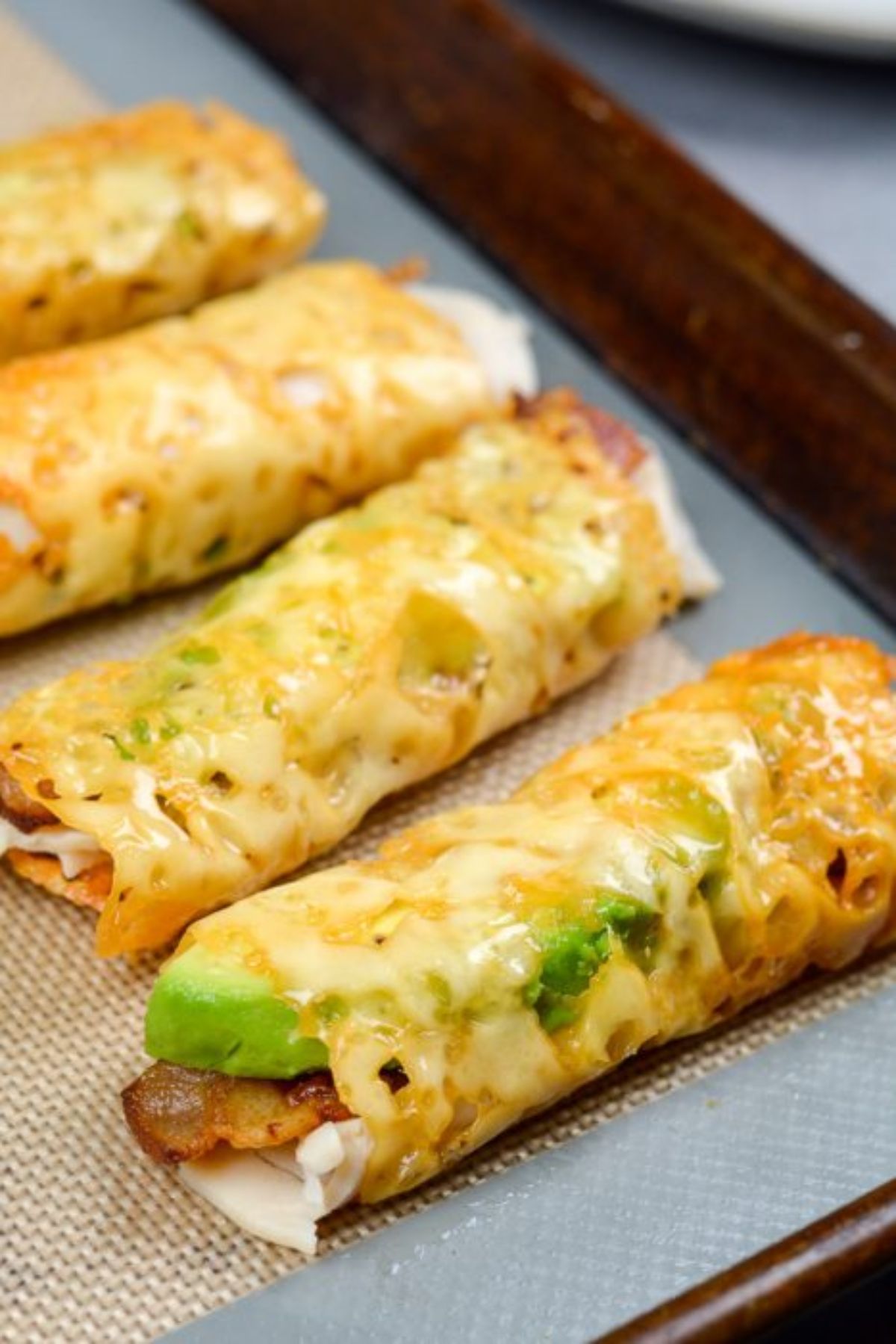 A partial shot of a lined baking tray with 4 taquitos on it. Avocado slices can be seen poking out