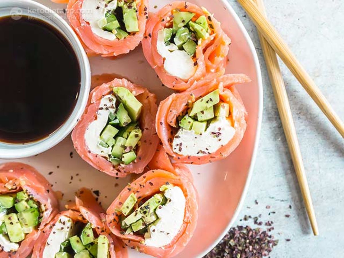 On a pink plate 7 salmon sushi rolls can be seen from above. To the left of them is a bowl of soy sauce and to the right 2 chopsticks