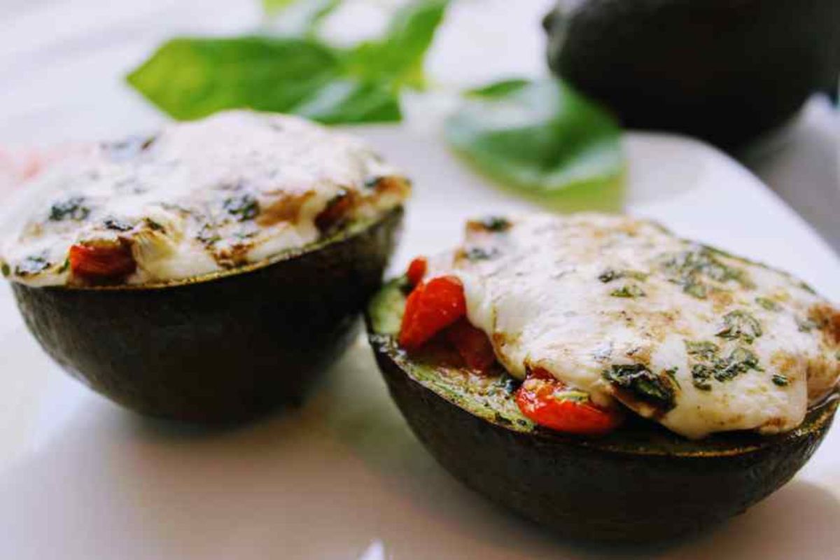 2 avocado halves are on a white surface. They are stuffed with vegetables and topped with melted cheese and herbs