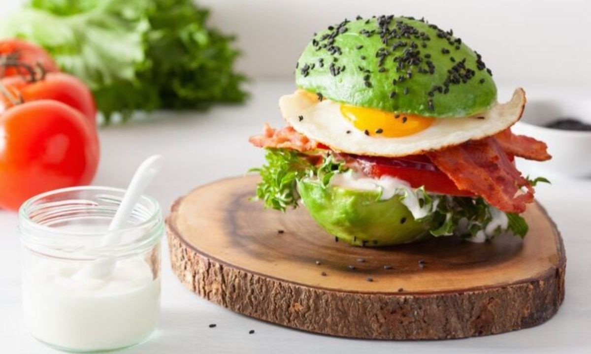 On a white countertop is a slice of tree trunk. On top of this is a peeled avocado, cut in half and acting as a burger bun around slices of bacon, a fried egg, and some lettuce leaves. To the left is a glass jar of sauce, tomatoes and a head of lettuce