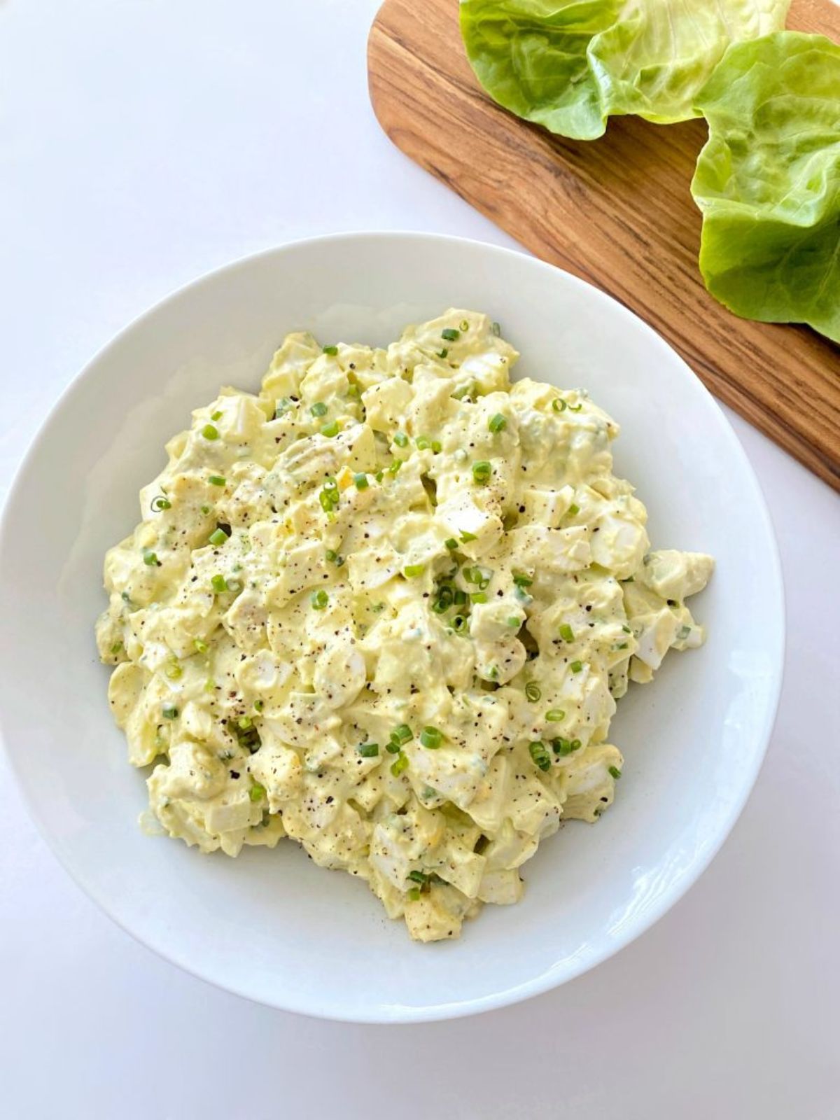 A white round bowl sits on a white counter holding a pile of avocado egg salad. To the right, just in shot, is the corner of a wooden chopping board with lettuce leaves on it