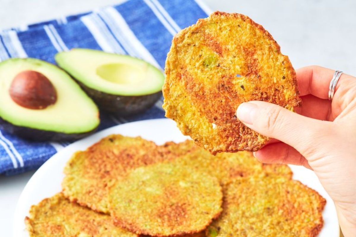 A hand holds an avocado chip in front of a plate of them. Behind is a blue and whote striped cloth with a halved avocado on it