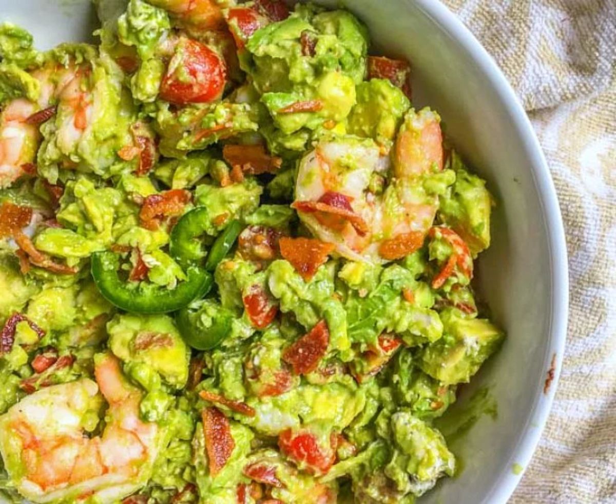 A white bowl filled with a creamy salad of peppers, shrimp, avocado, bacon bits, tomatoes and lettuce