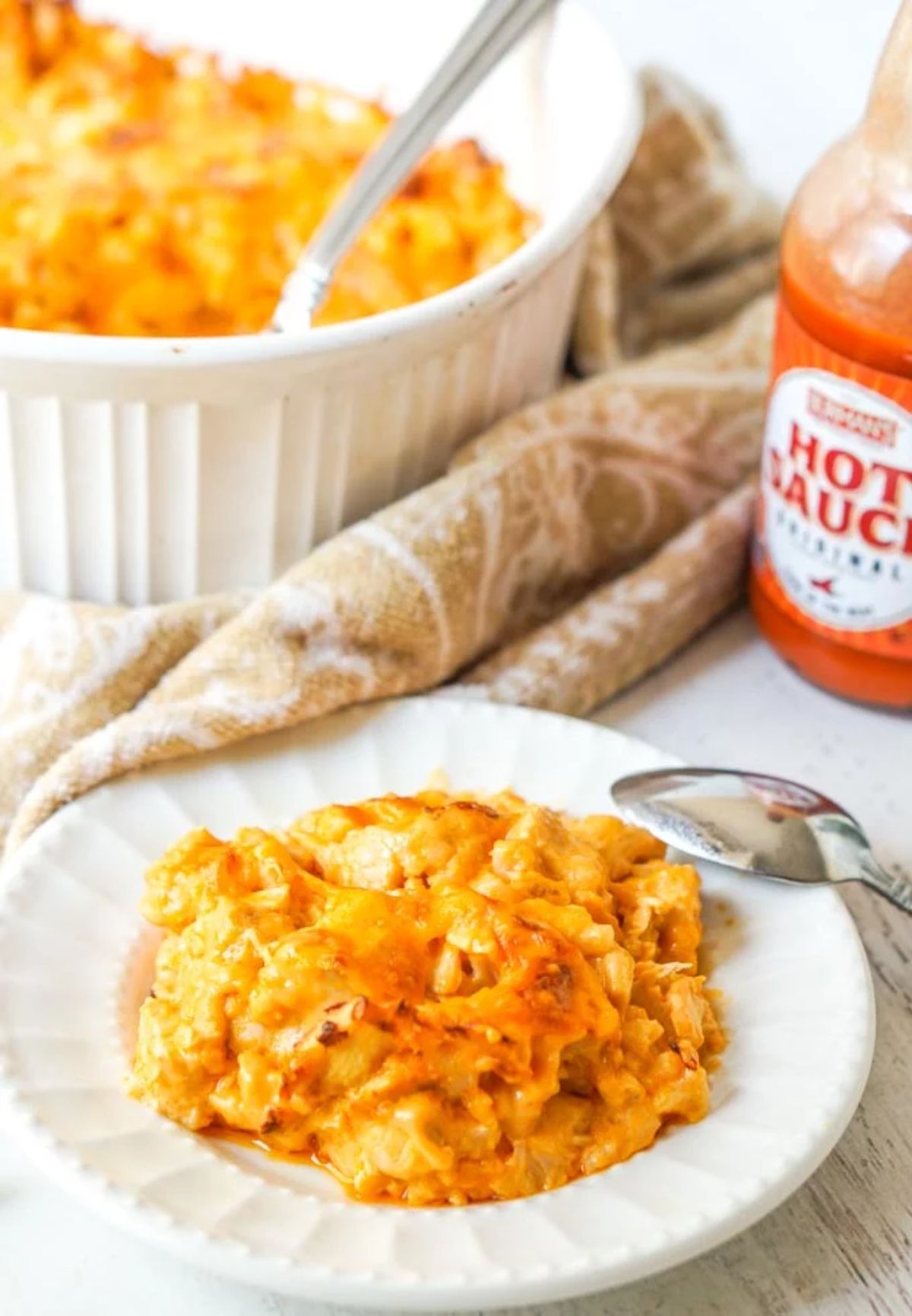 in teh back left of the shot is a white deep casserole dish with cheesey casserole in it. On the right is a bottle of hot sauce. In the foreground is a white plate with a portion of casserole on it and a spoon resting on the edge