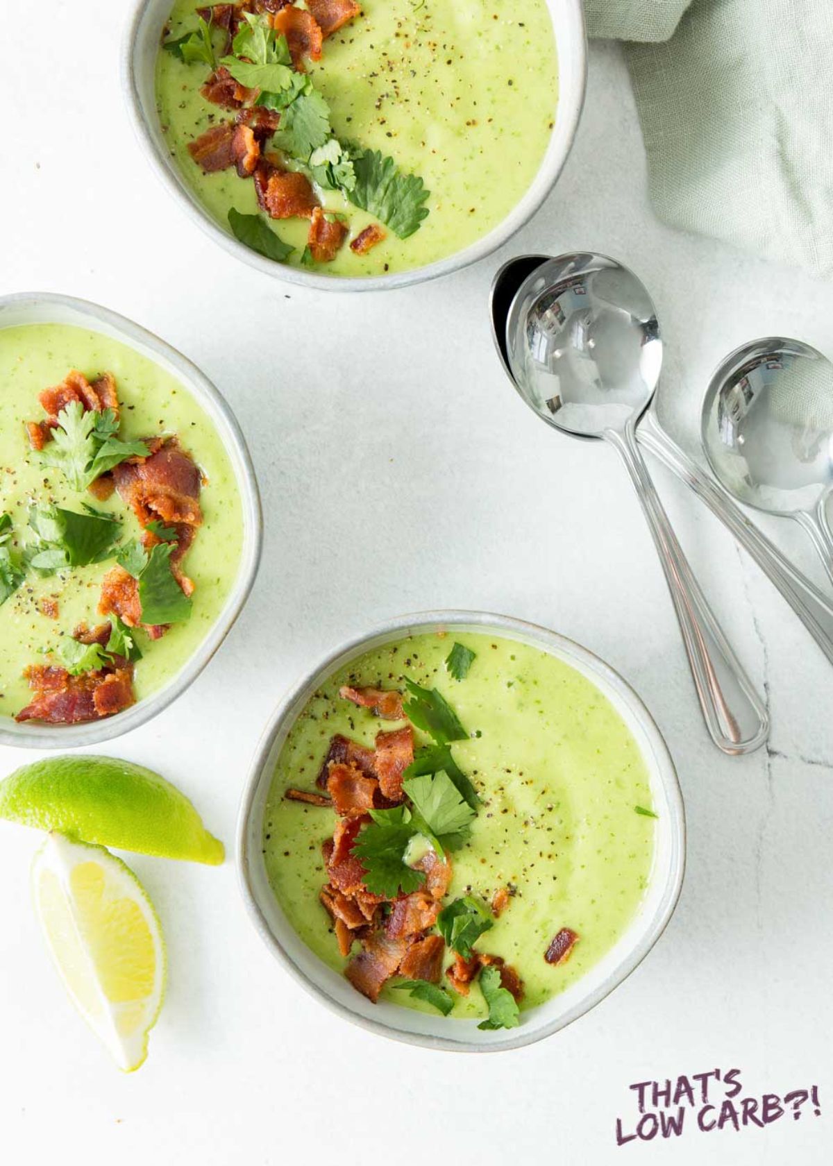 A top view of 3 bowls of avocado soup on a white counter top. They are topped with bacon bits, and chopped herbs. Lime wedges and soup spoons are next to them
