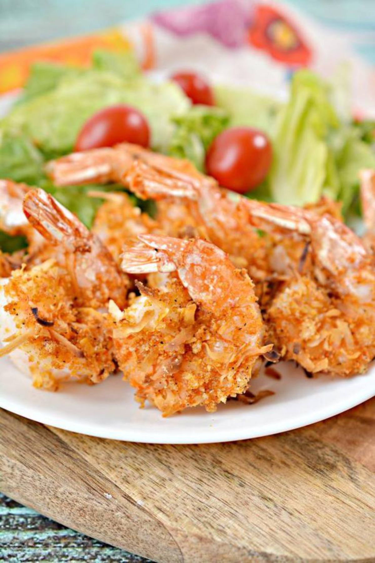 On a partially seen wooden board is a whote plate fll of coconut shrimp in front of a lettuce and cherry tomato salad