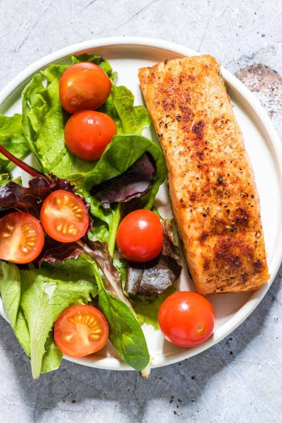 A round white plate holds a salmon fillet and a salad of green leaves and halved cherry tomatoes