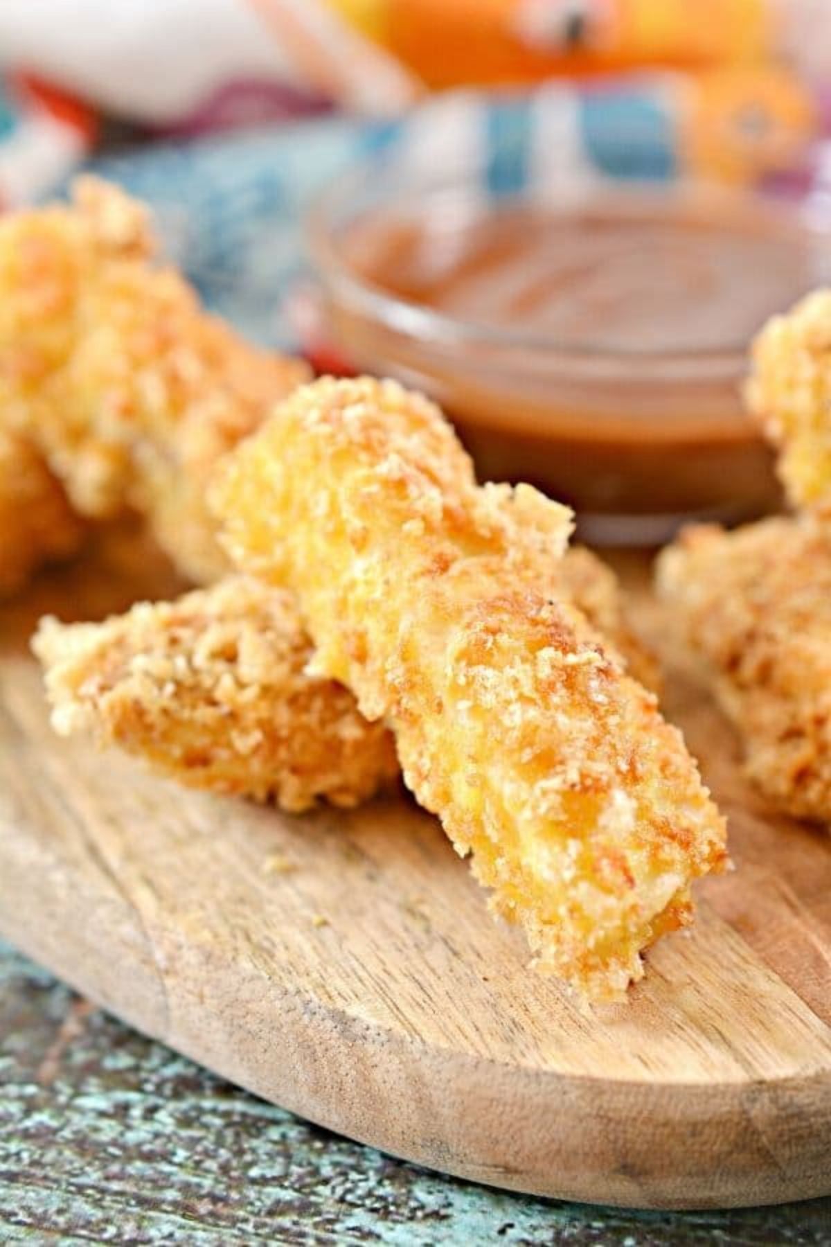 A partial shot of a wooden board with fish sticks stacked on it Behind is a blurred galss bowl with a red dip in it