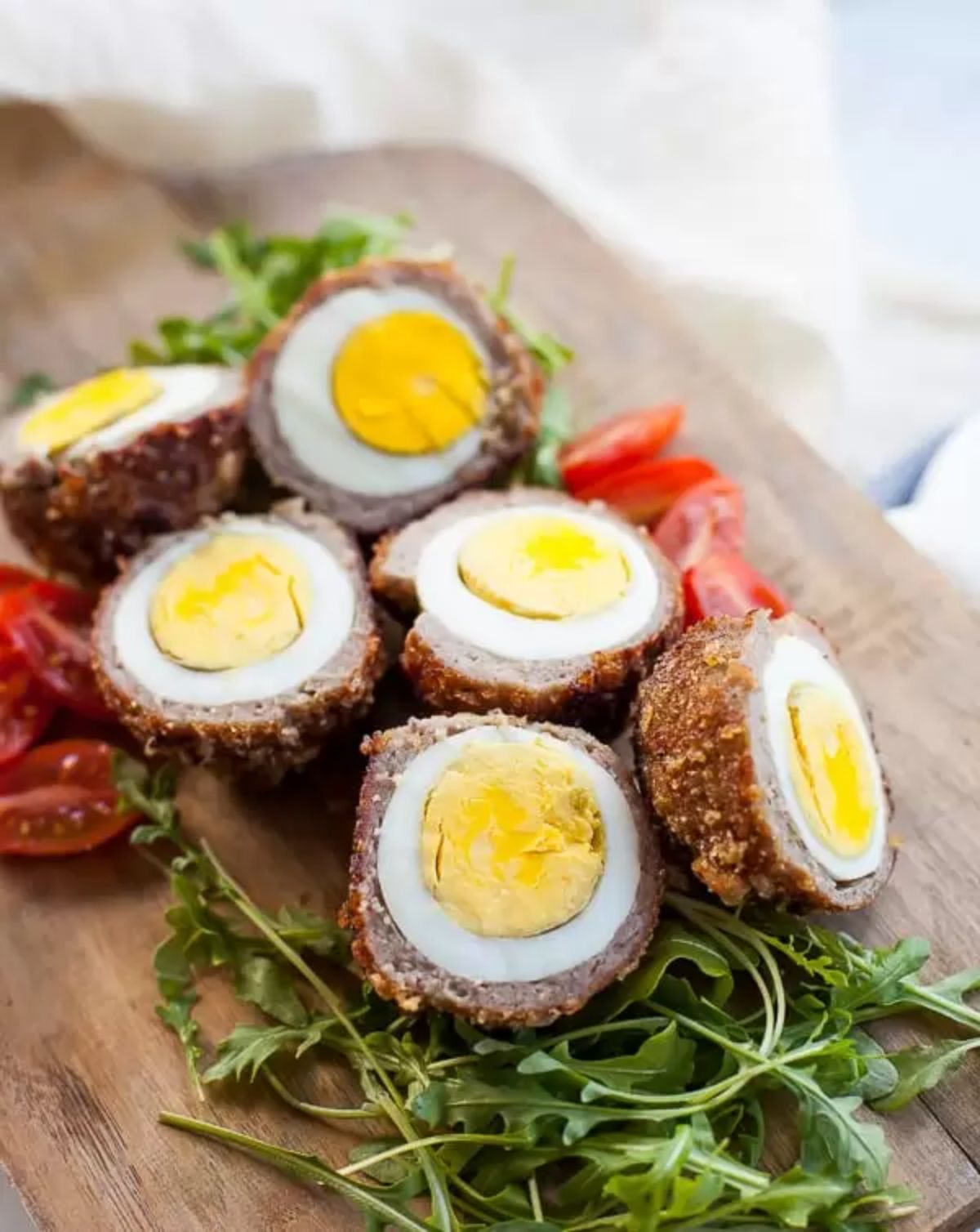 A wooden chopping board has 3 halved scotch eggs on a bed of arugala leaves and tomatoes