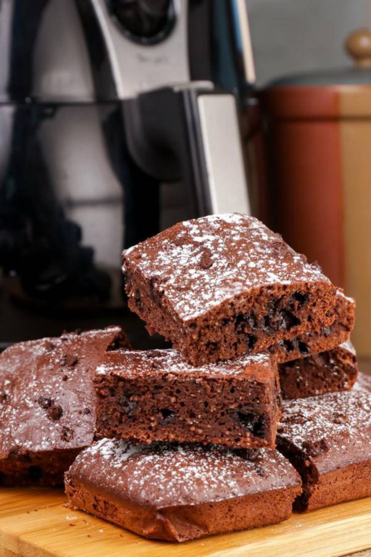 In front of an air fryer on a wooden board is a pile of chocolate brownies