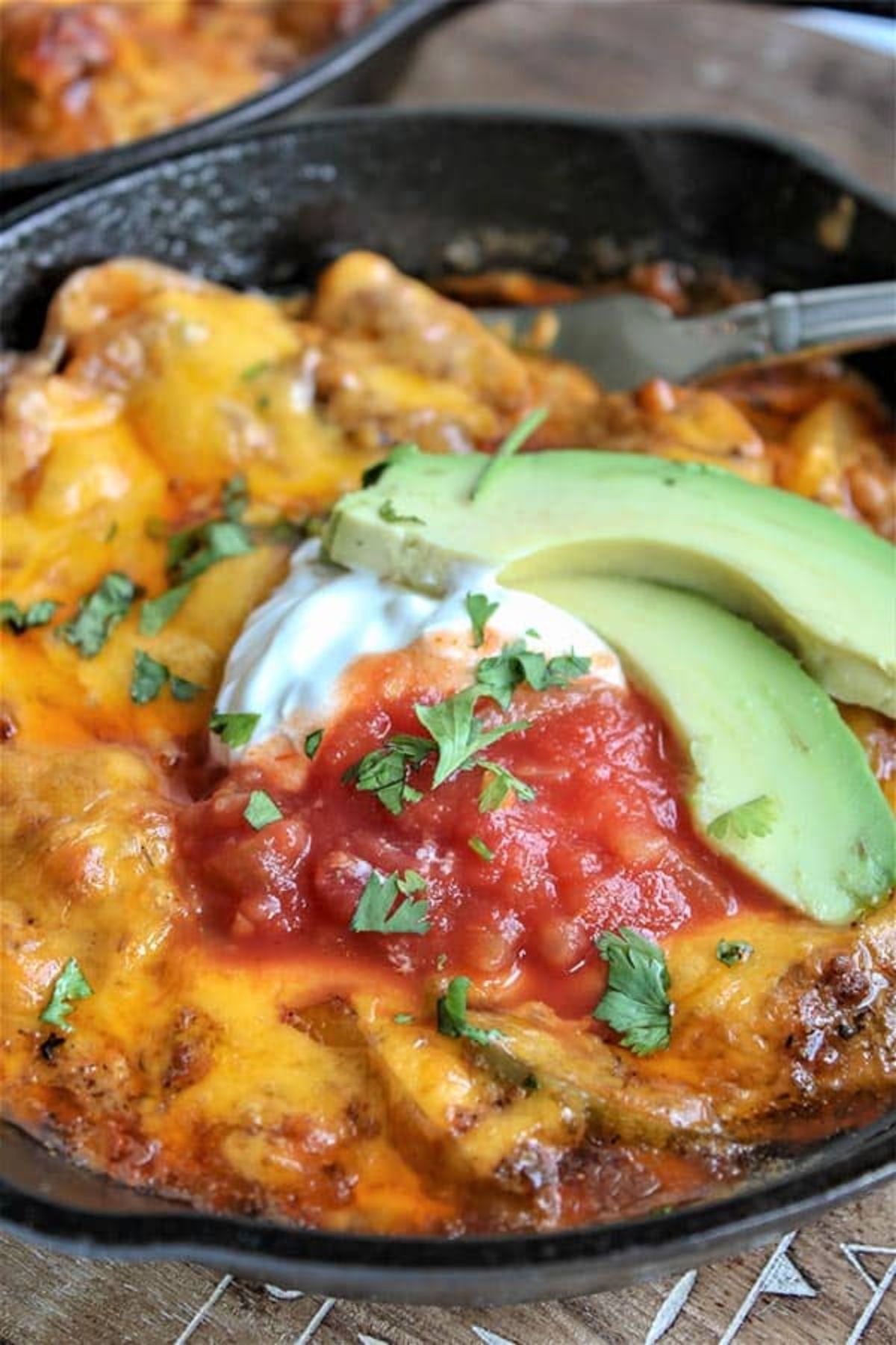 In an iron skillet is a cheesey casserole, garnsihed with cliced avocado, salsa and sour cream