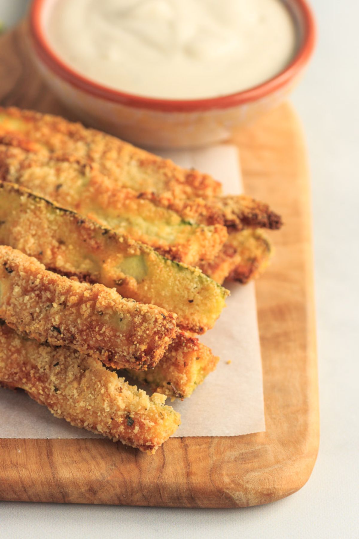 A partial shot of a wooden chopping board with zucchini fries in a pile at the front. Behind is a pottery bowl full of white dip