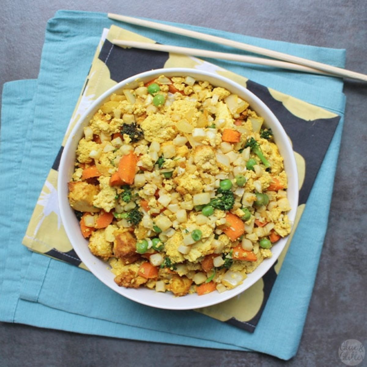 On a folded light blue cloth and flowery napkin is a round white owl of cauliflower rice with chopped vegetables/ Two chopsticks sit above it