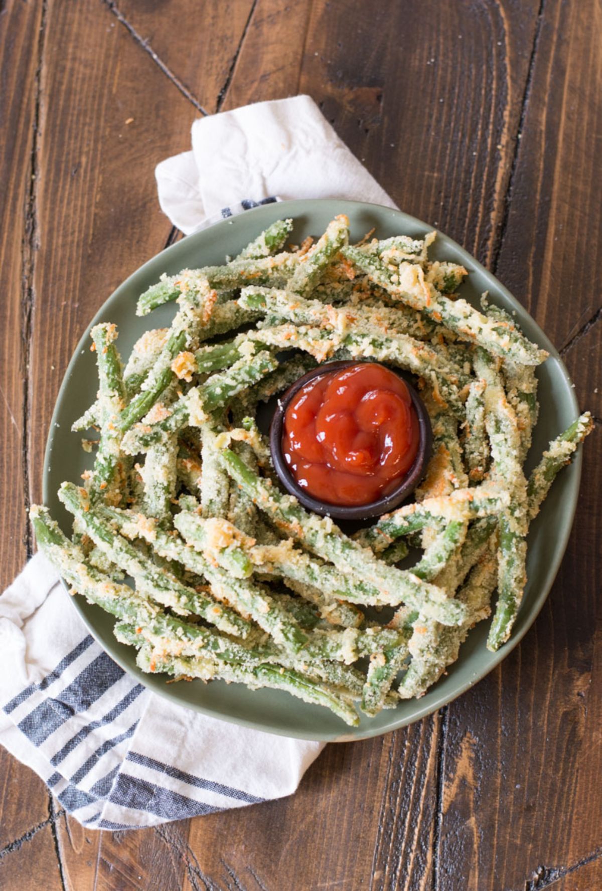 On a dark wooden table is a white and blue cloth with a green bowl on top. A pile of fried green beans surround a pot of red dip