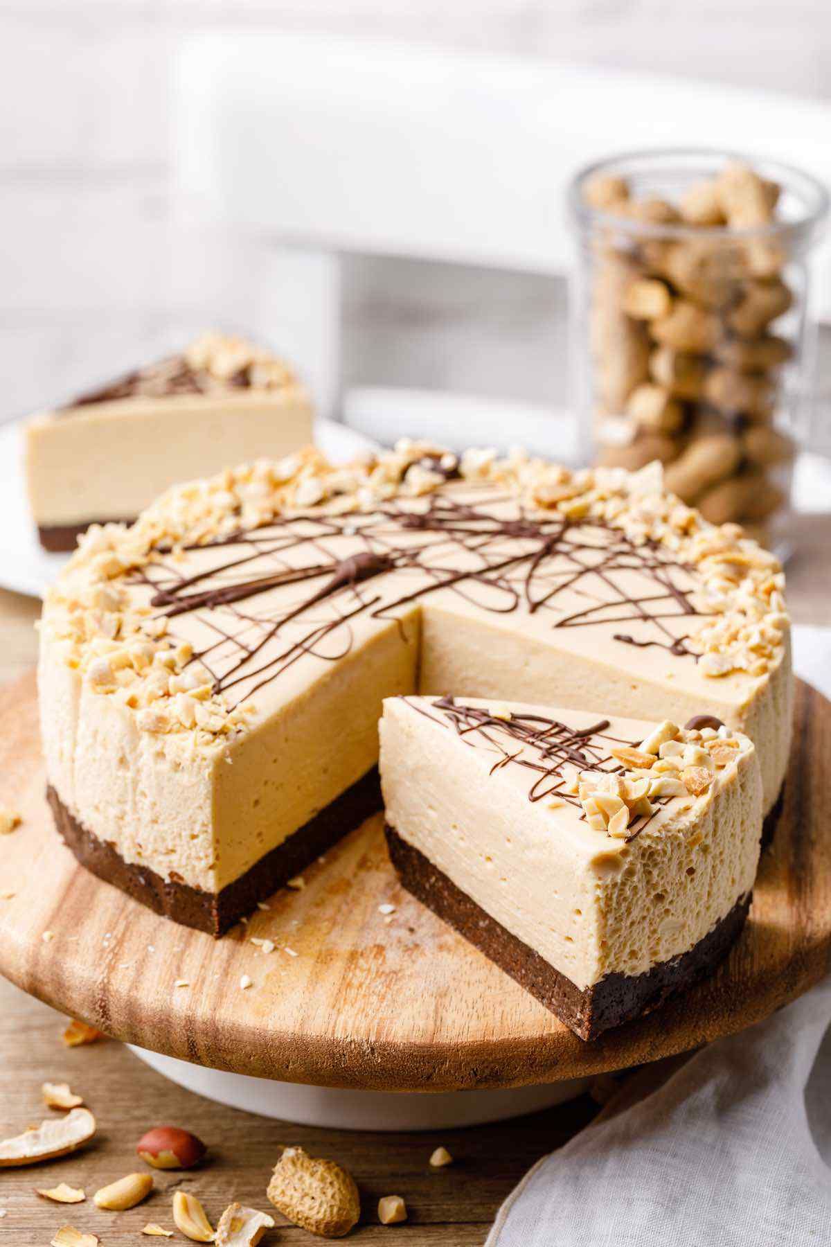 On a wooden lazy Susan is a cheesecake with a dark chocolate base and caramel colored filling. One slice has been cut out. There are peanuts scattered over the table
