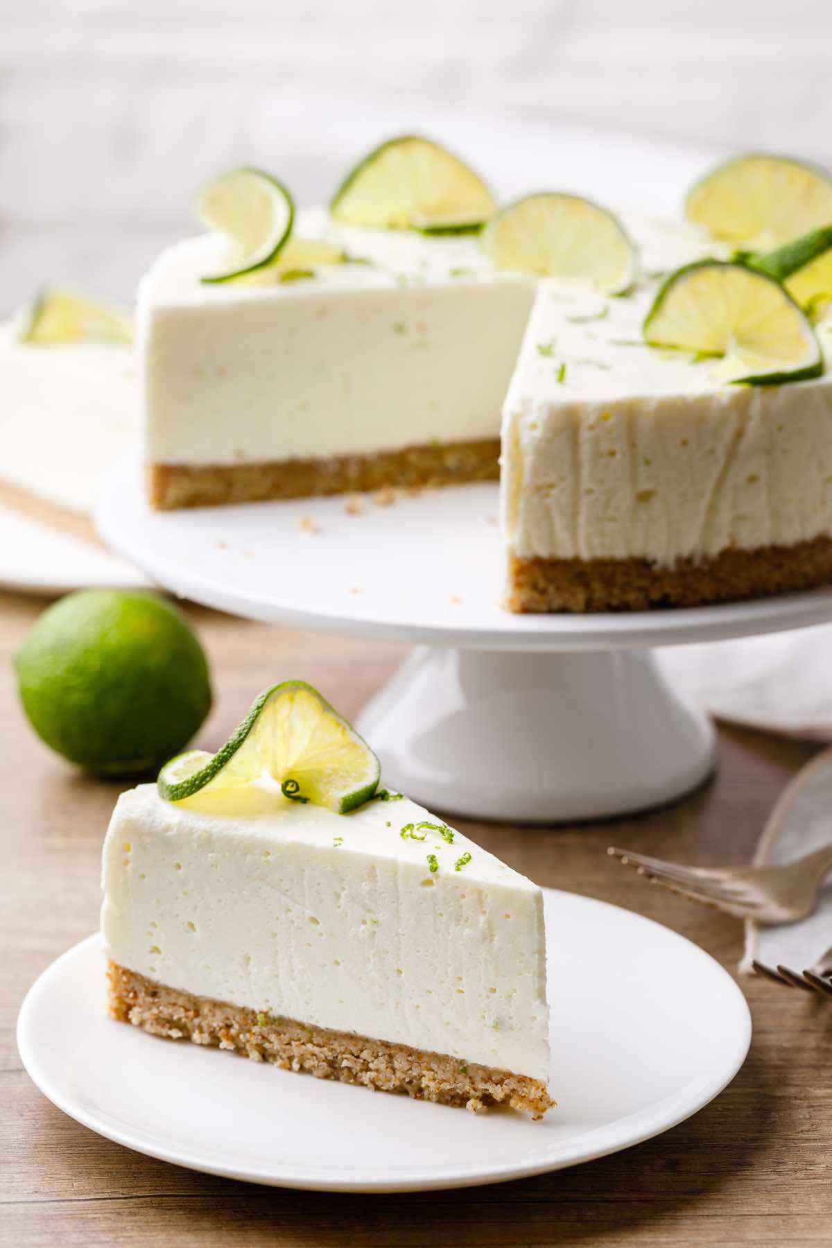 on a cake plate is a lime cheesecake with a portion taken out. on a plate below is one slice of the cheesecake with lime twist on top