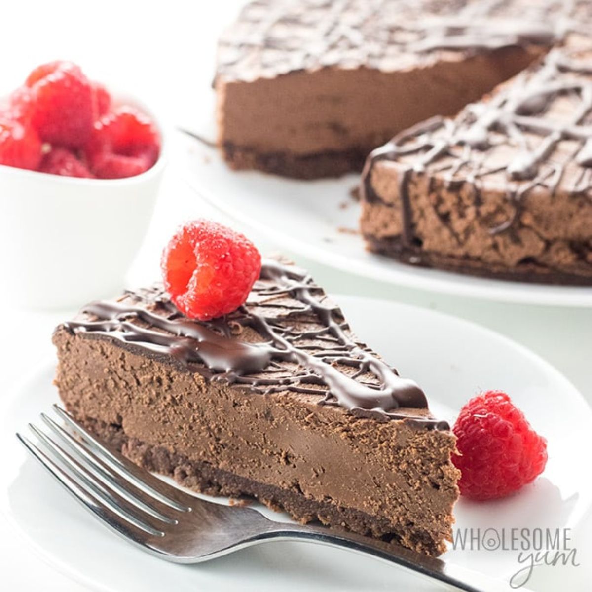 on a cakestand int he back of the photo is a chocolate cheesecake with chocolate drizzle. The cut out slice is on a white plate in front and is topped with raspberries