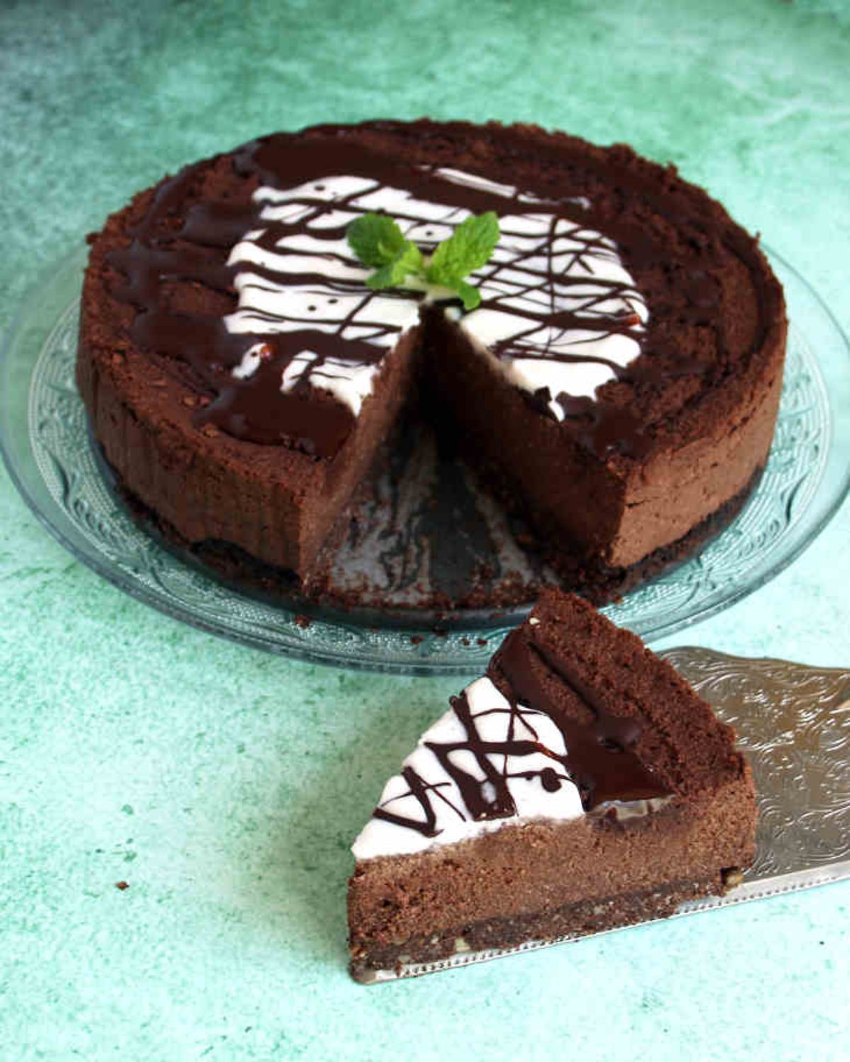 a dark chocolate cheesecake sits on a glass cake stand. It is toped with chocolate drizzle and some mint leaves. On a cake slice is the msising slice of cake