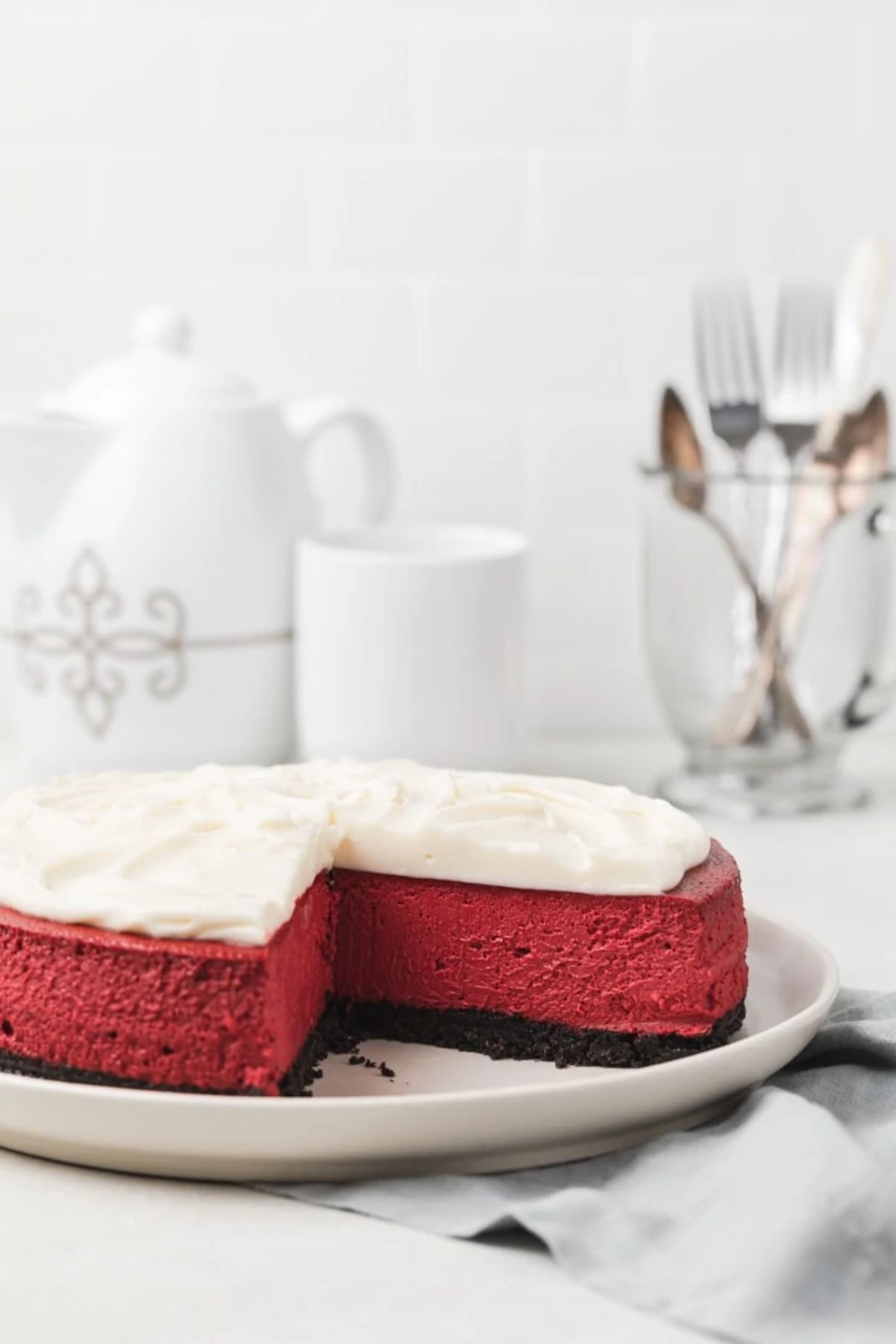 In front of a white china tea set is a red velvet cheeskecake with cream topping anfd a missingslice