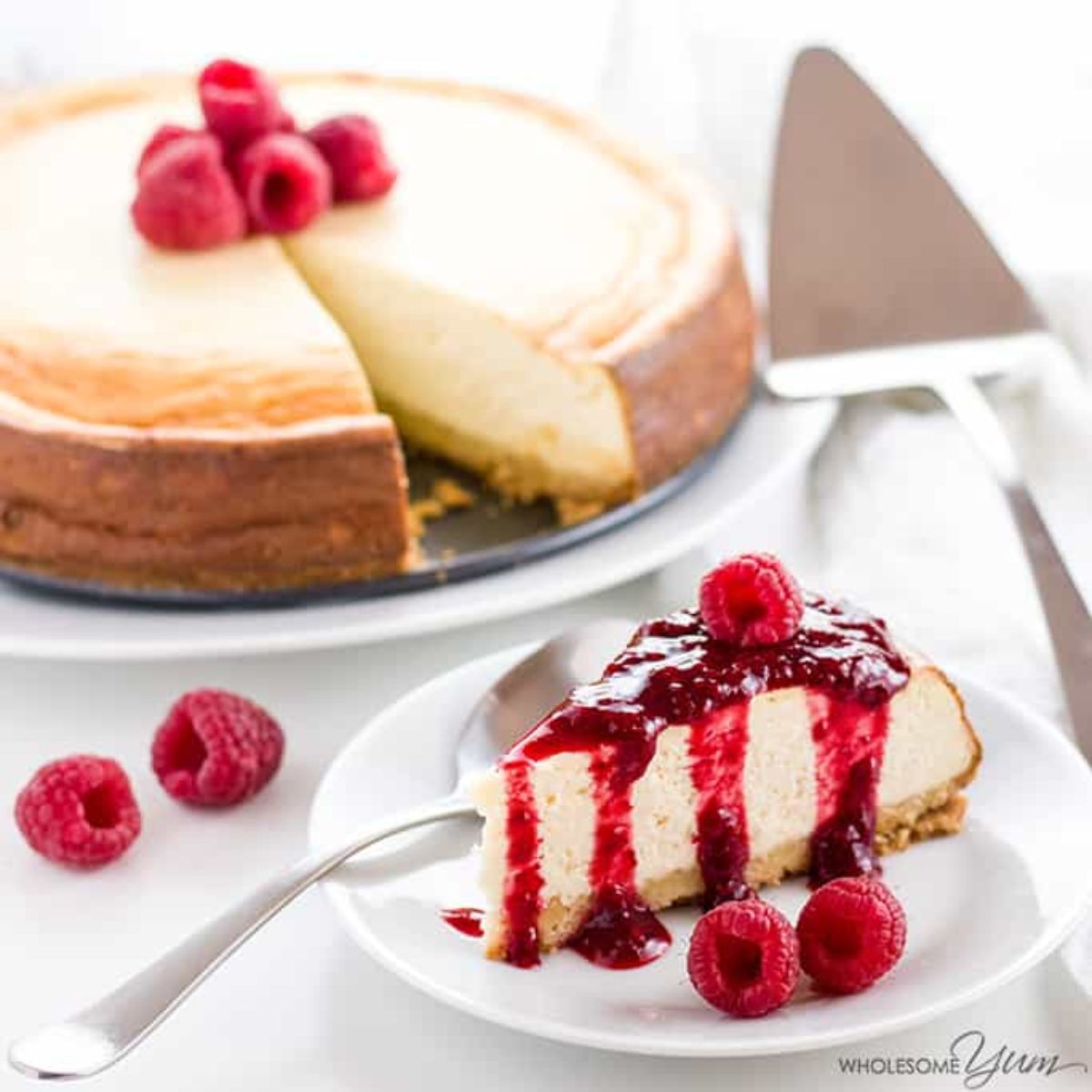 on a plate is a baked cheesecake with raspberries in the center. Below it is one slice of the cheesecake with raspberry coulis dirzzled over it. A cake slice os on the righ tof the photo