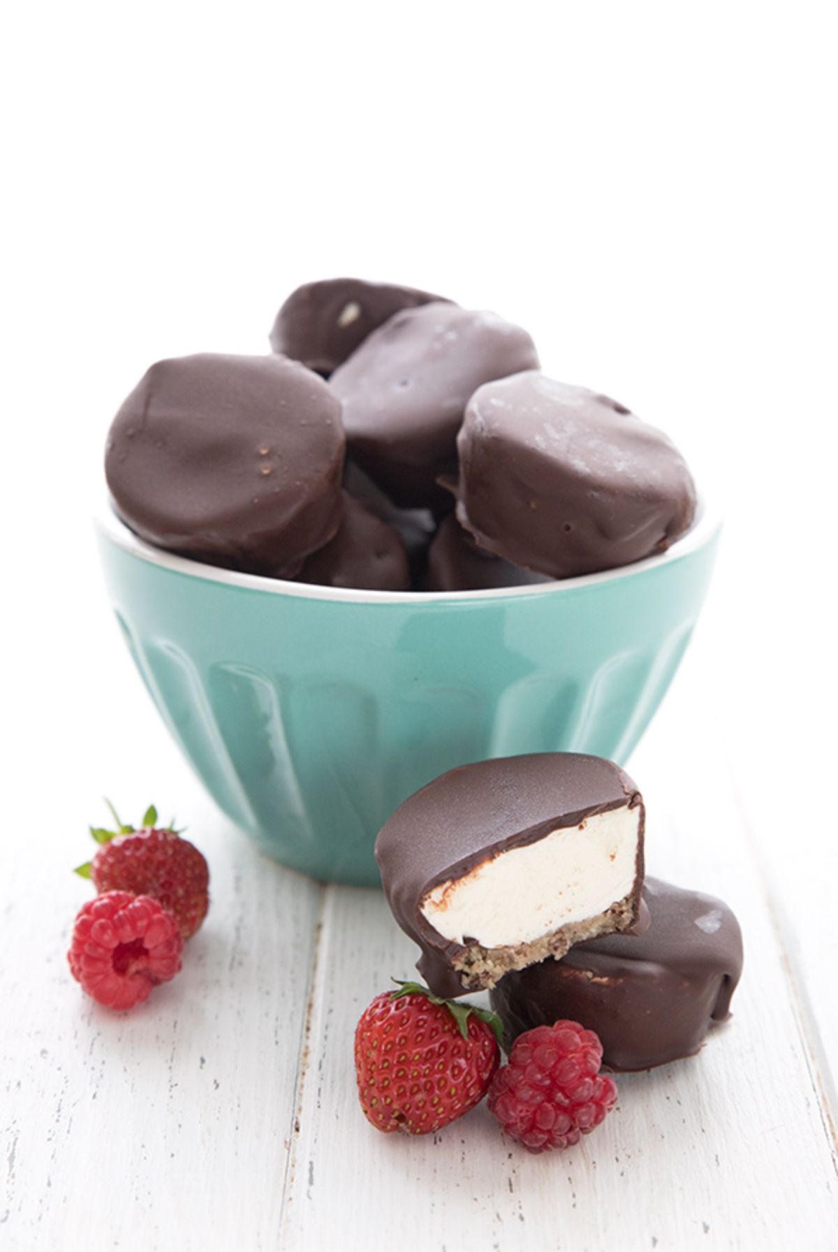 A ligth green bowl hold a pile of chocolate covered cheesecake bites. In front are 2 of the bites and a pile of raspberries