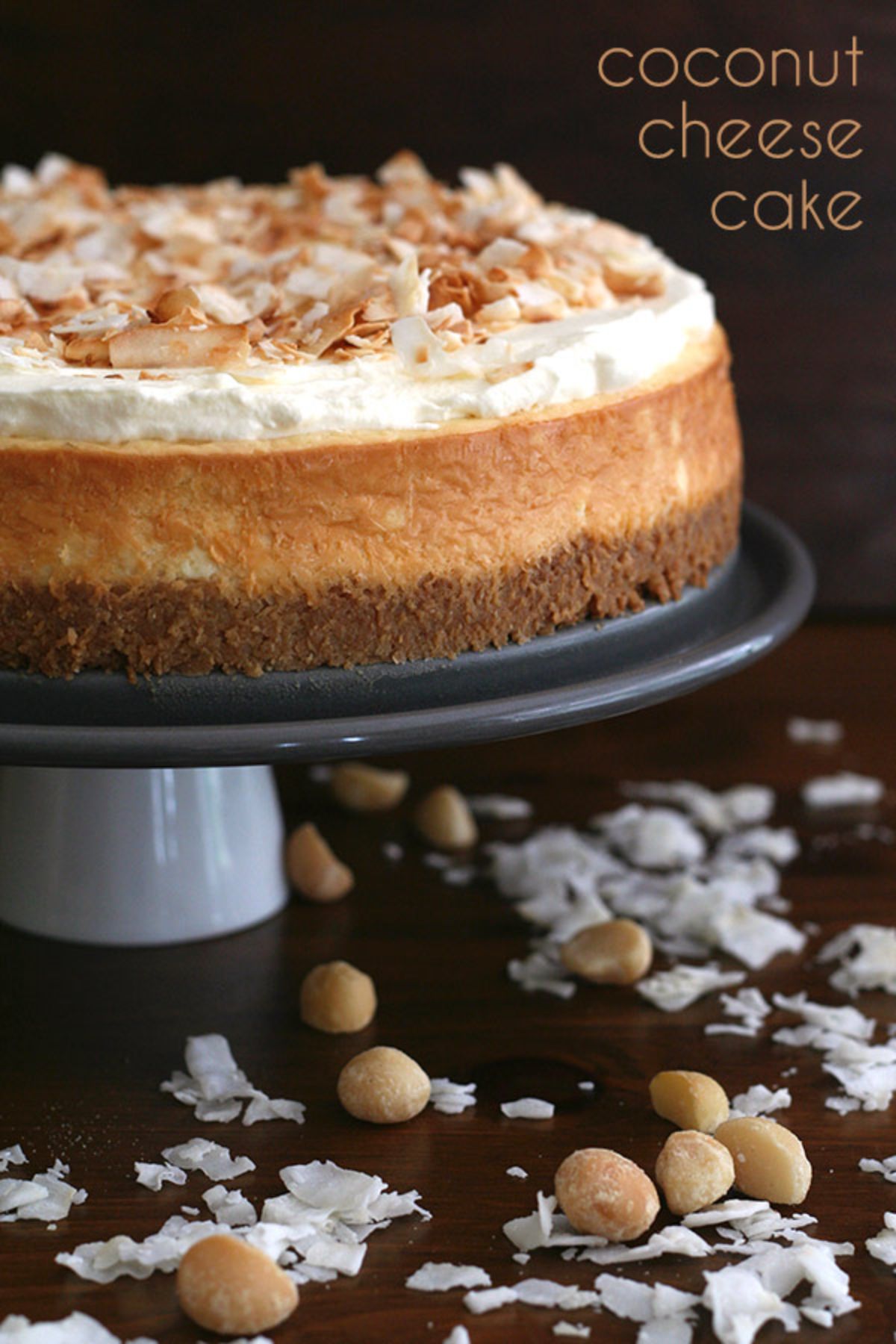 On a drak background a 3 layered cheesecake sits on a ckae stand sprinknled with flaked almonds