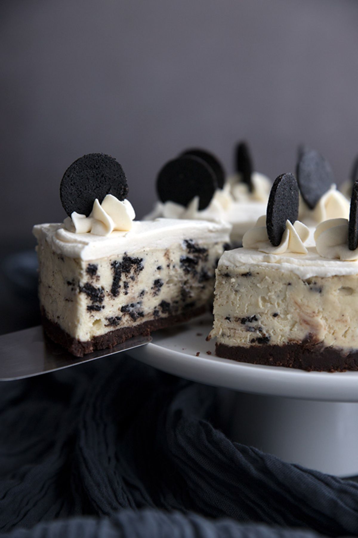 On a cake stand is a cookies and cream cheesecake. On top are rosettes of cream with oreos stuck in the to. A slice is being moved on top of a cake slice