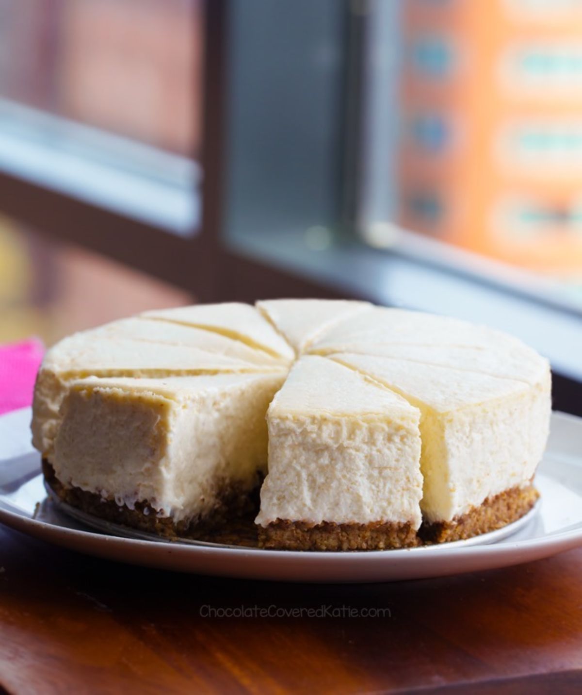 A plain cheesecake sits on a cake stand in front of a window. It has been cut into 8 slices