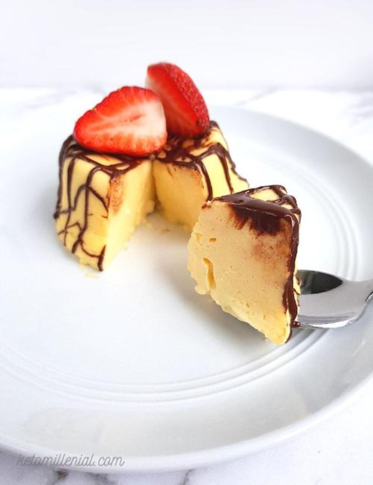 In the middle of a white plate is a round pudding laced over with chocolate and two strawberries on top. a spoon has taken a chunk out of it