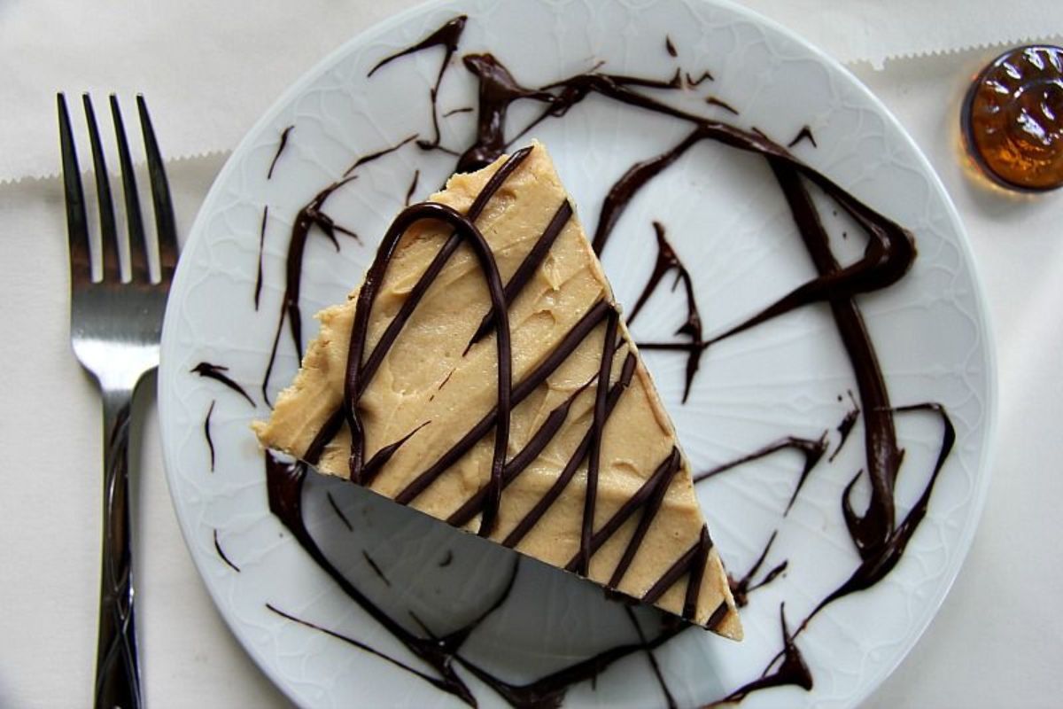 on a white plate is a triangular slice of vanilla cheesecake with chocolate drizzled over it