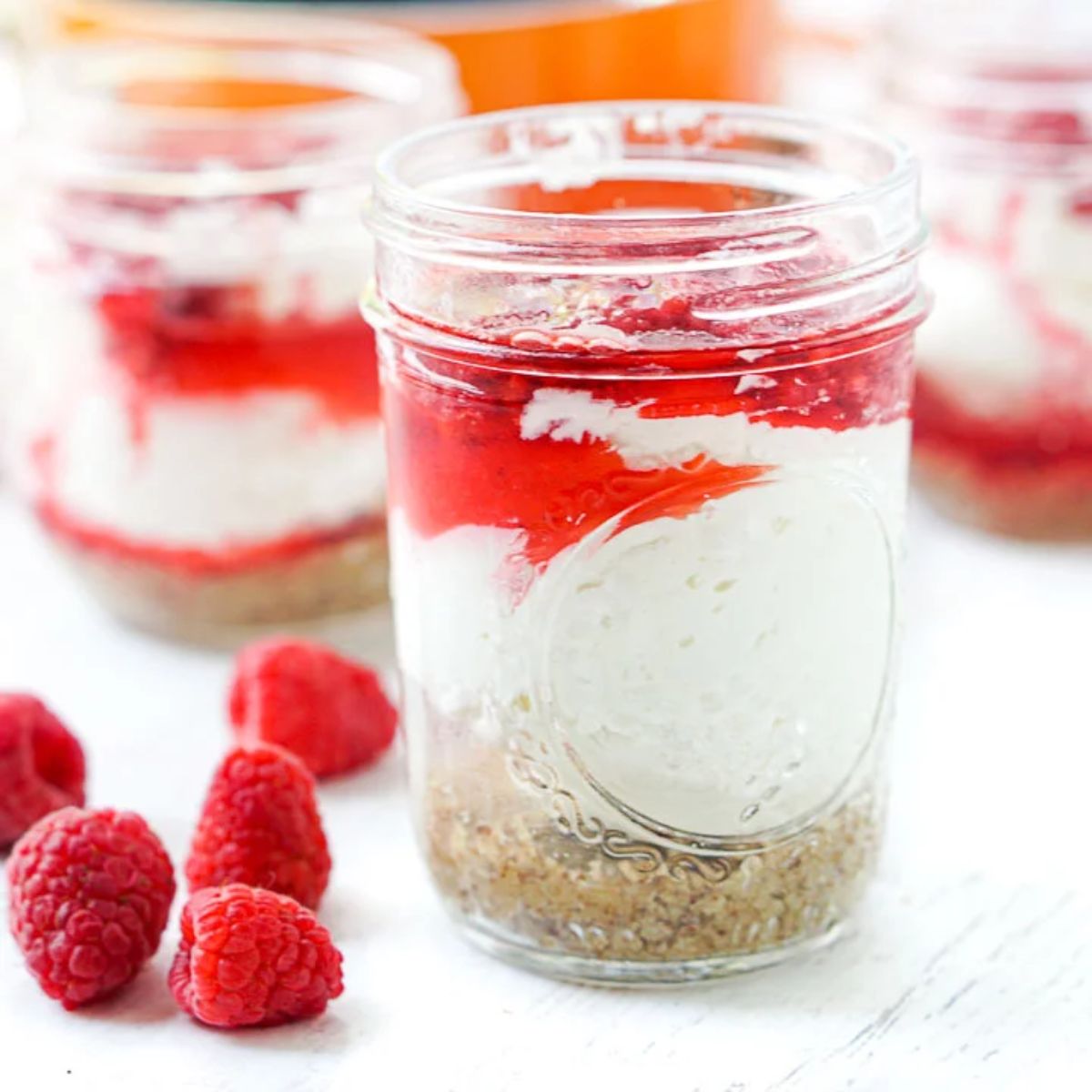 3 small glass jars filled with layers of biscuit, cream and red furit coulis. A pile of raspberries sits in front