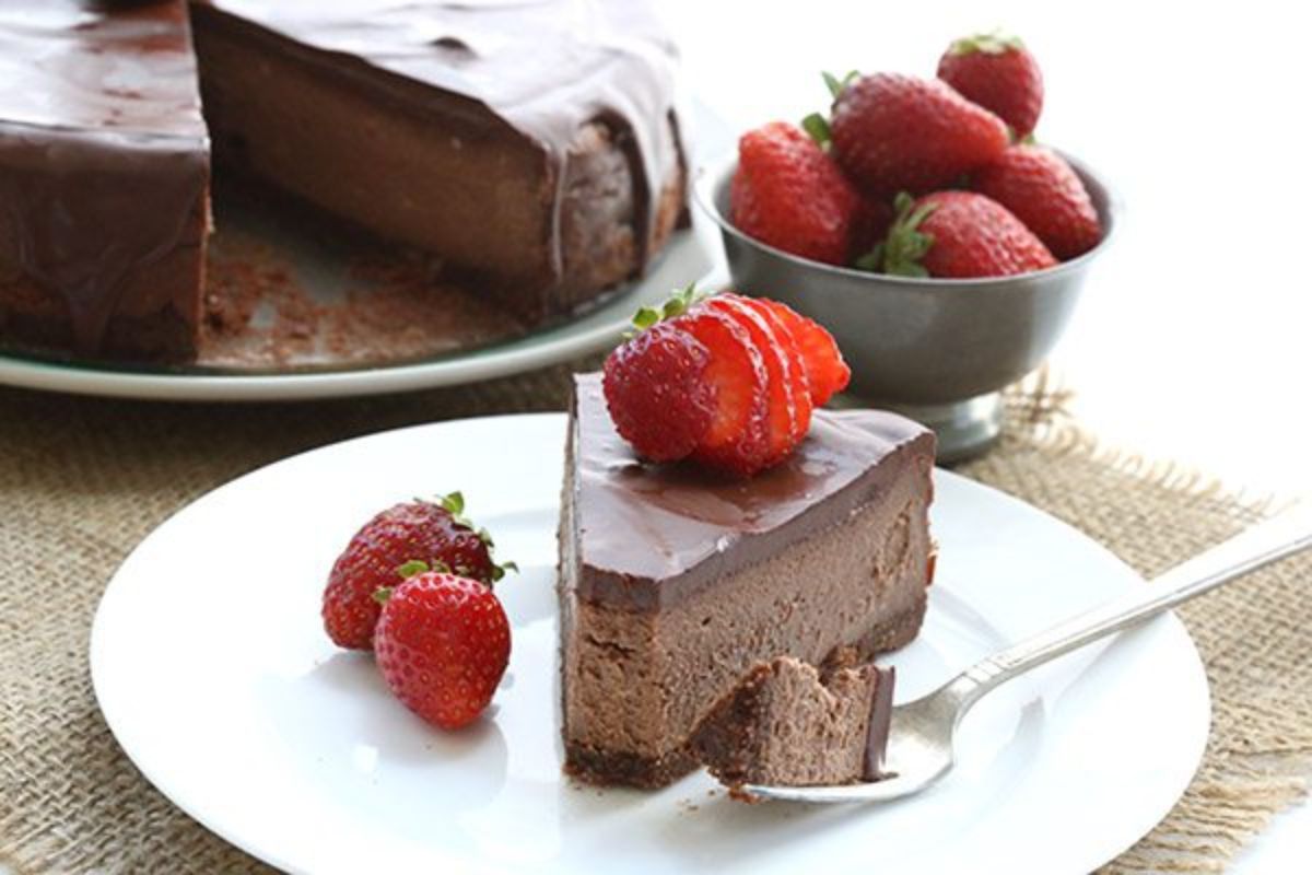 In the back of the photo is a chocolate cheesecake with a cdarker chocolate top and bottom with a slice cut out. The slice sits in frot on a white plate with strawberries on top and behind it is a bowl of strawberries