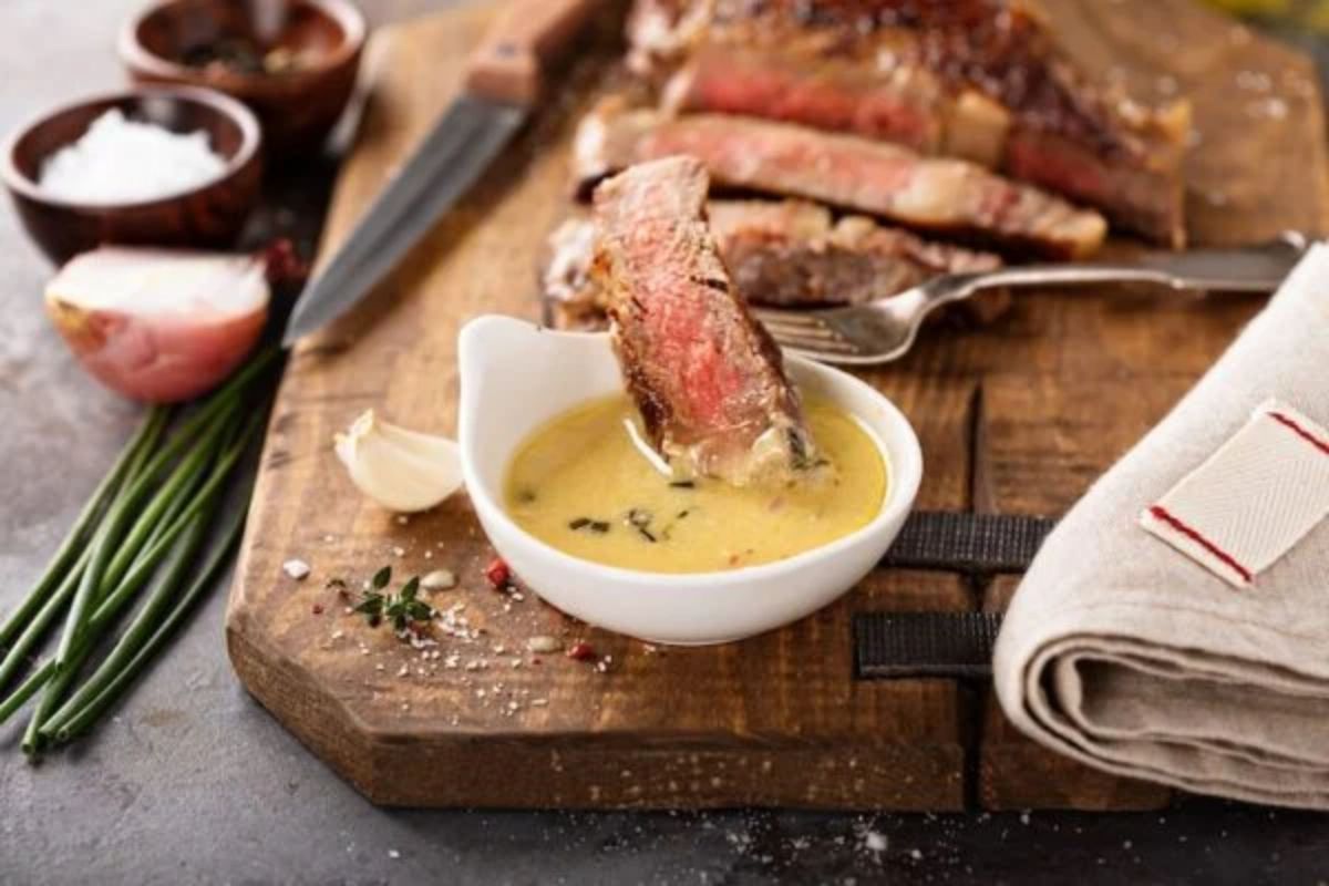 A chopping board sits on a gray s stone surface. On it is a sliced steak and a white pot full of yellow sauce. A fork with a piece of steak on the end is resting over the sauce. Also in shot are wooden bowls with salt and pepper, half an oinon, scallions, and a hessian cloth
