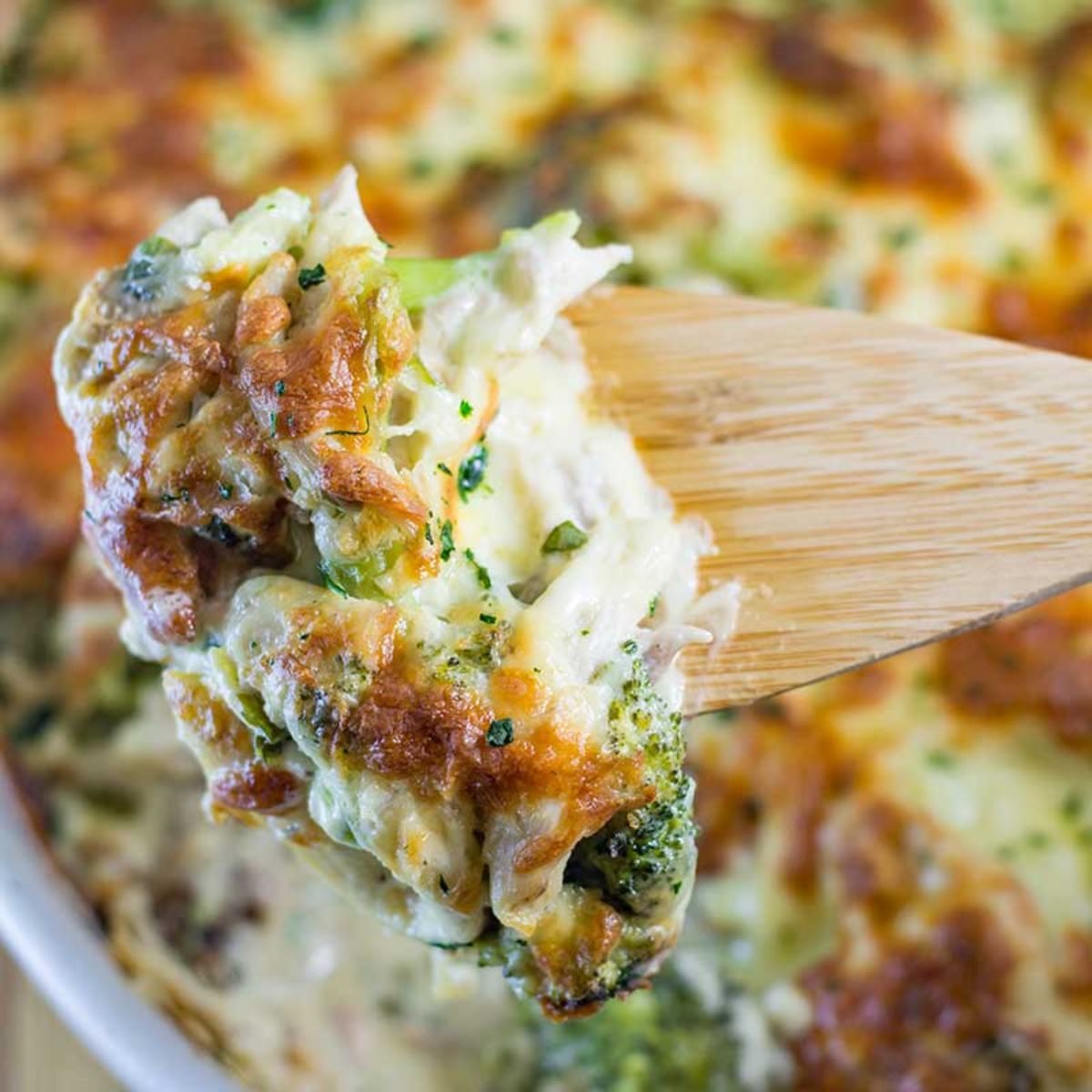 a blurred casserole can be bseen in the background. In the foreground a wooden spoon lifts a portion out of the dish