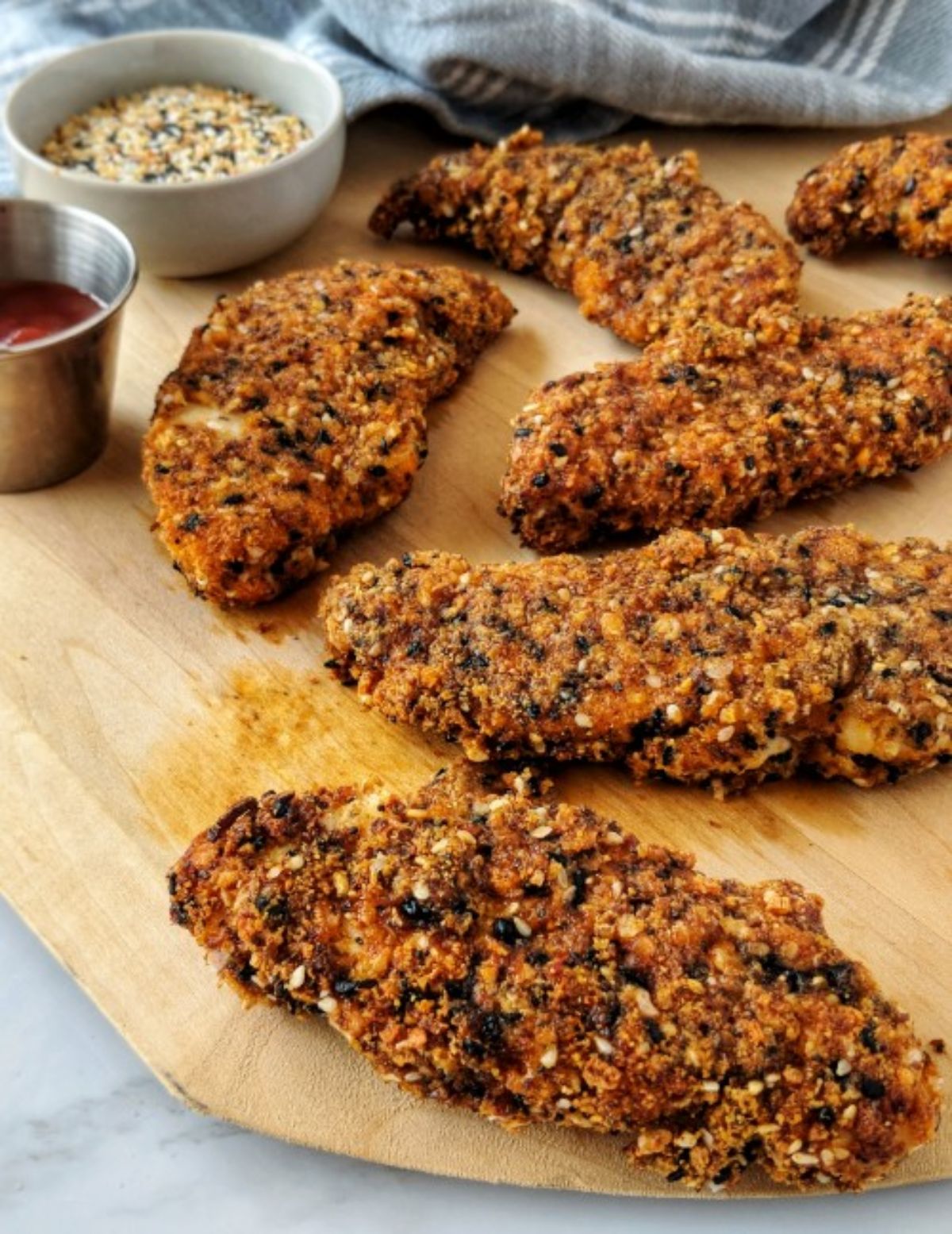 On a wooden board are seasoned chicken tenders with a white pot of seasoning and a metal pot of red sauce