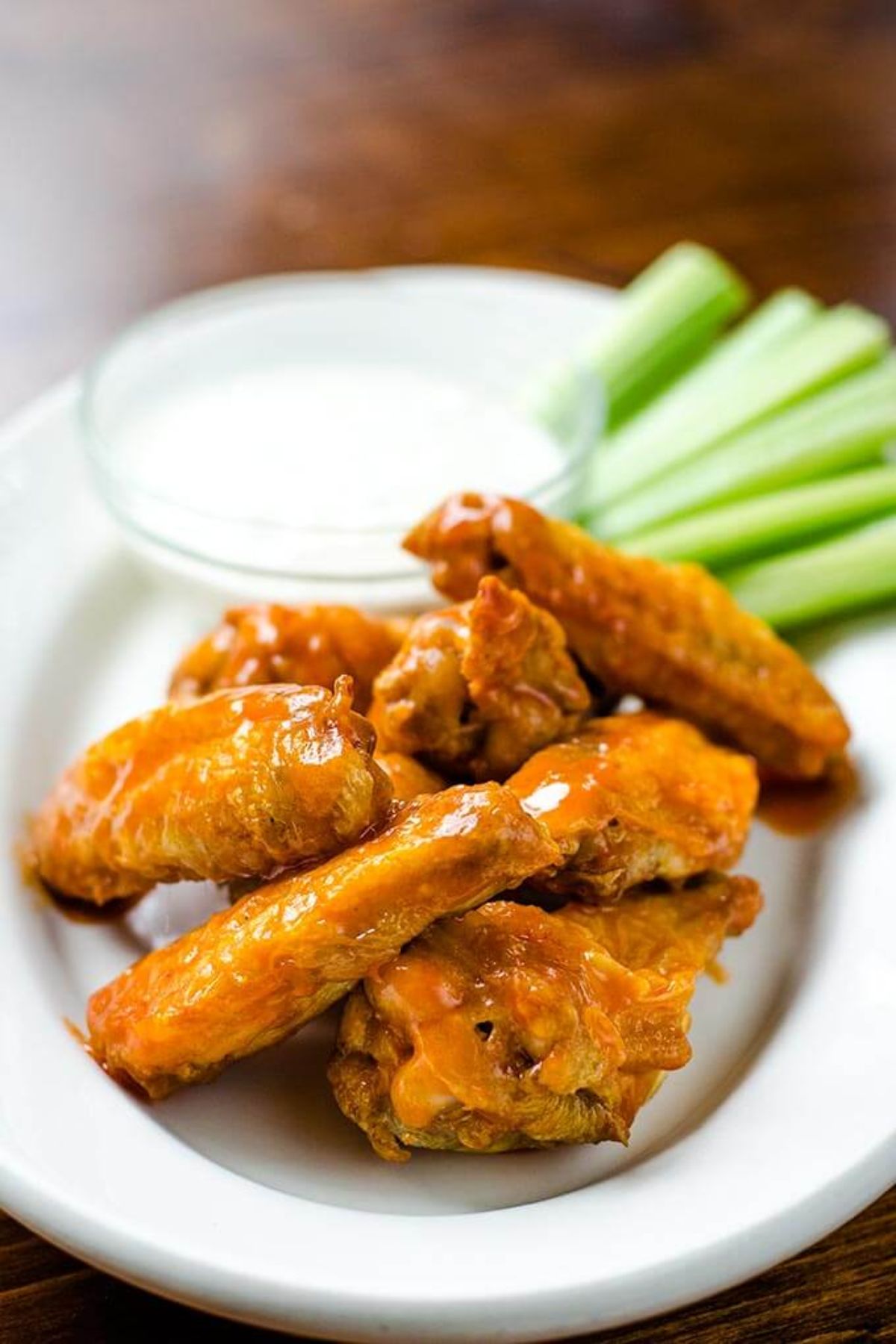 A small oval white plate has a pile of chicken wings on it, with a glass ot of white sauce and celery sticks in the background