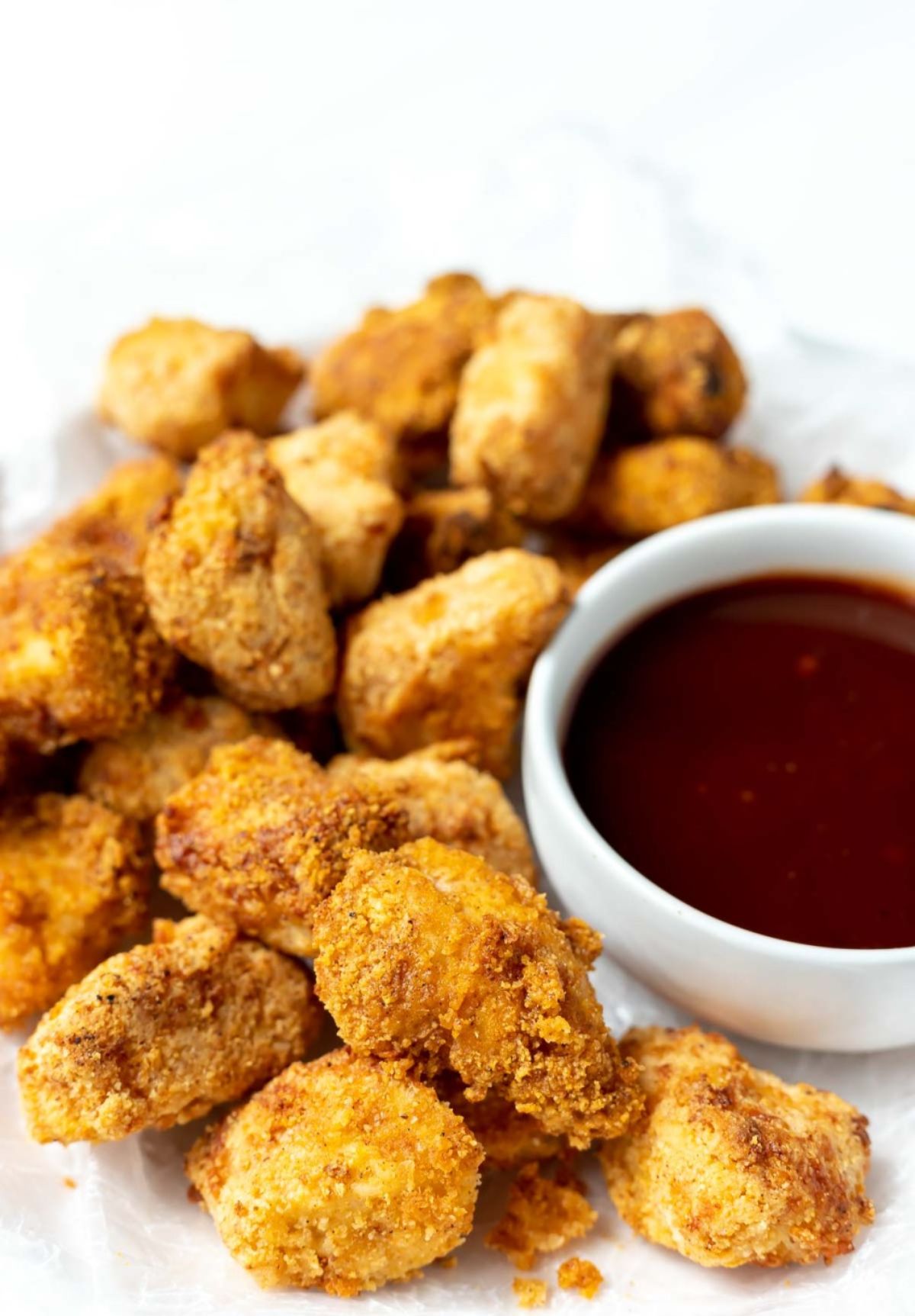 On a white plate is a pile of chicken nuggets with a white pot of red sauce to the right